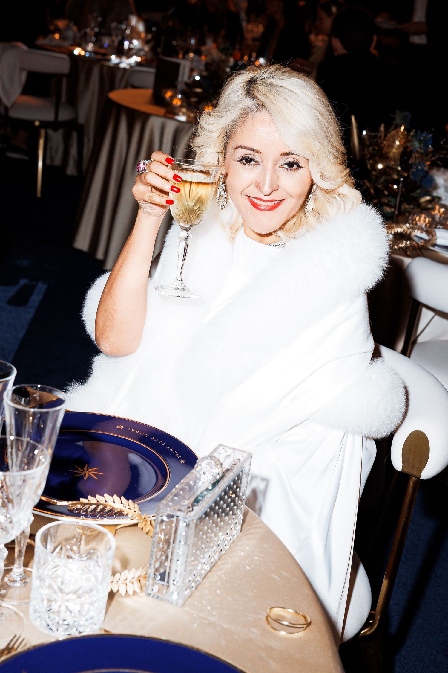 A person in a white fur coat sits at an elegant dining table with crystal glassware and raises their champagne glass.