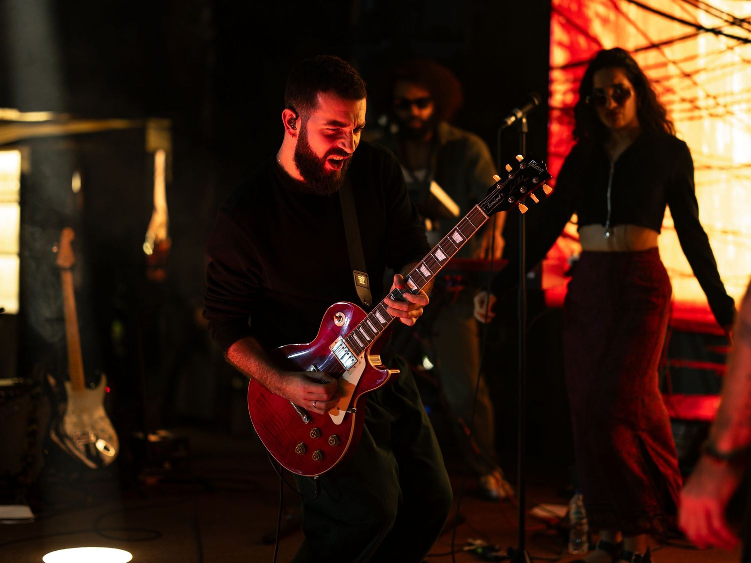 A guitarist performs on stage in a dimly lit venue with warm red lighting creating a moody atmosphere.
