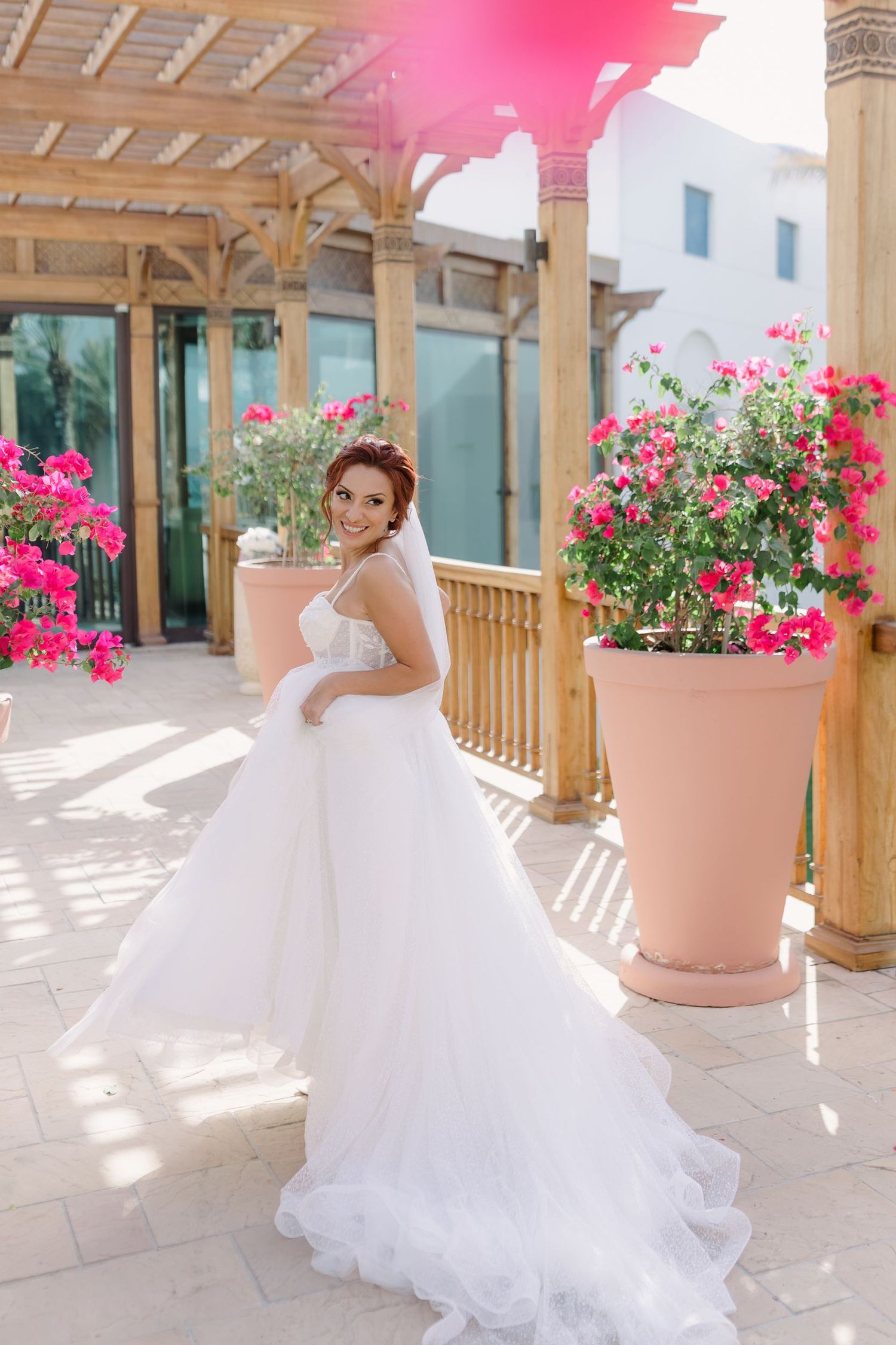 A white ballgown twirls on a sunny terrace with pink flowers and wooden beams.