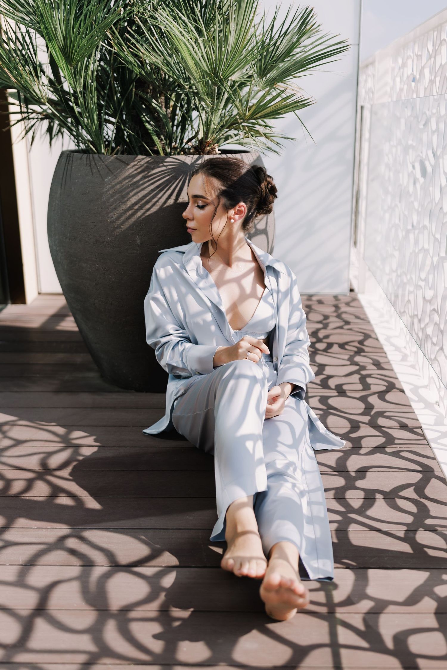 Fashion photo of model in white loungewear sitting by tropical plant with dramatic shadows.
