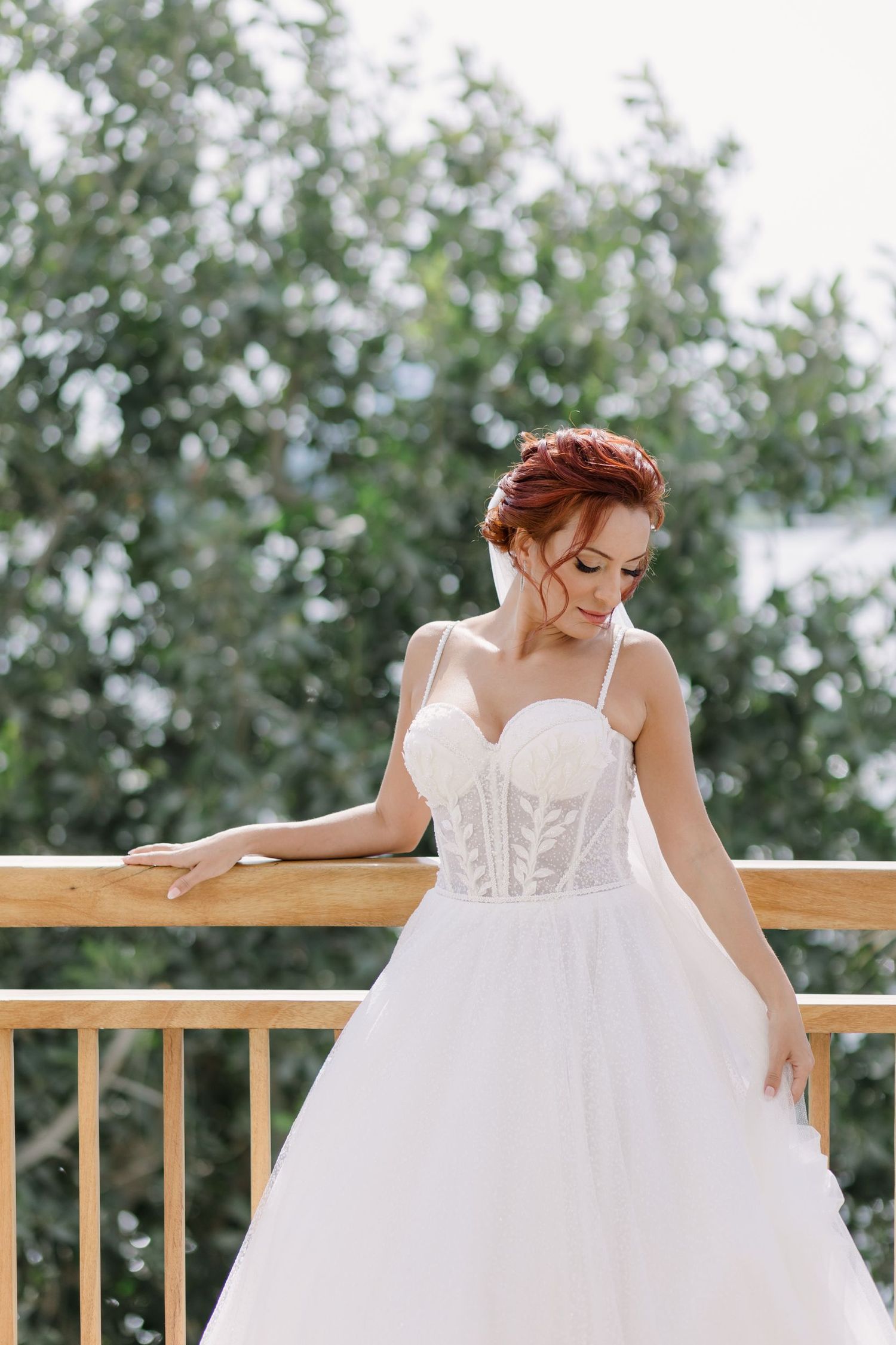 A flowing white wedding dress with a corset bodice is shown against a wooden deck railing with greenery in the background.