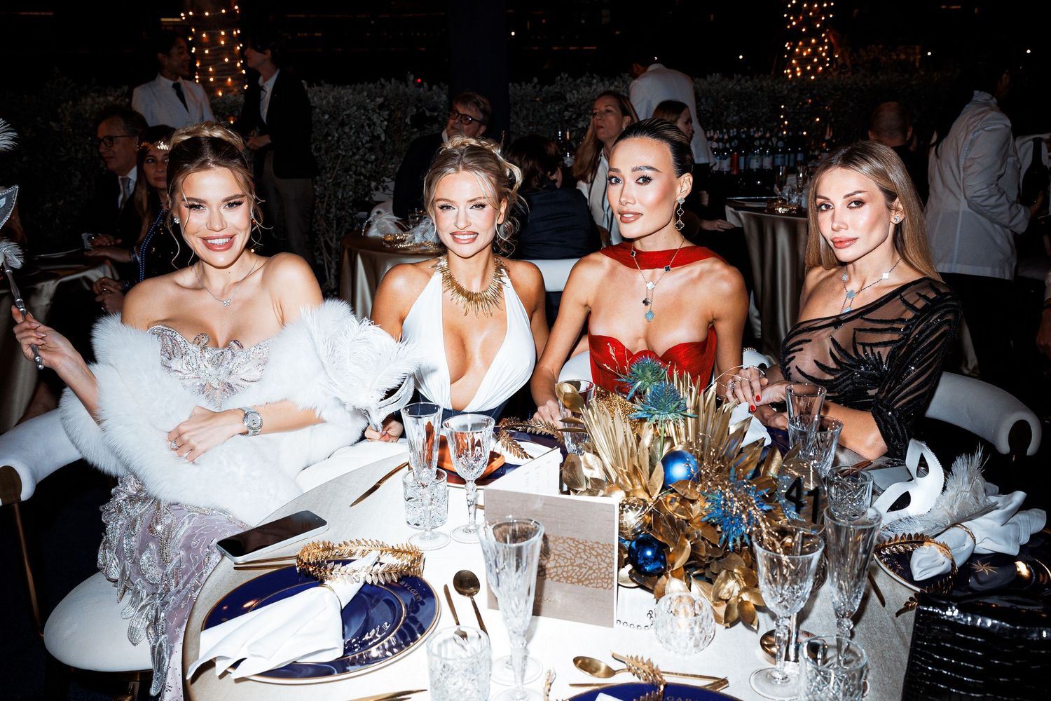 A glamorous group sits at an elegant dinner table decorated with masks and party decor for a formal masquerade event.