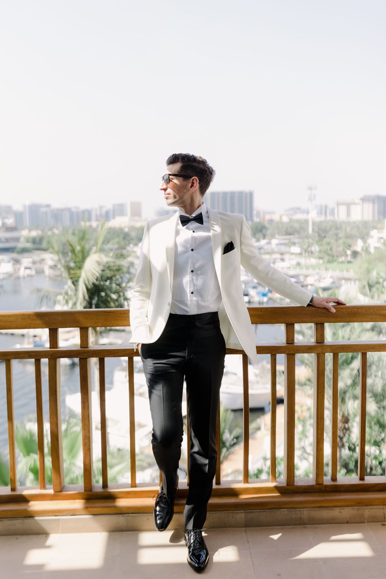 Stylish groom in white dinner jacket poses on balcony overlooking city skyline.