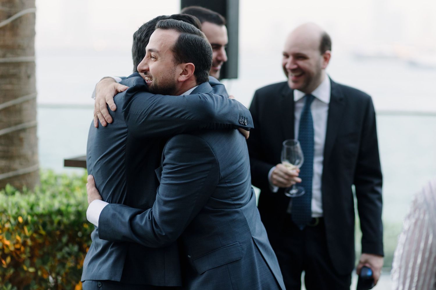 Two men in dark suits share an emotional embrace while another looks on at an outdoor event.