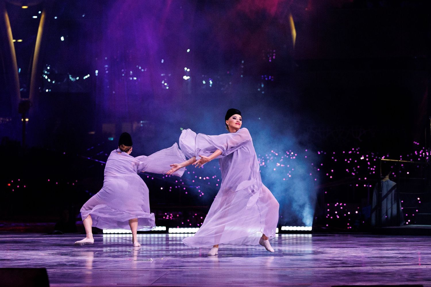 Dancers in flowing white costumes performing under purple and blue stage lights.