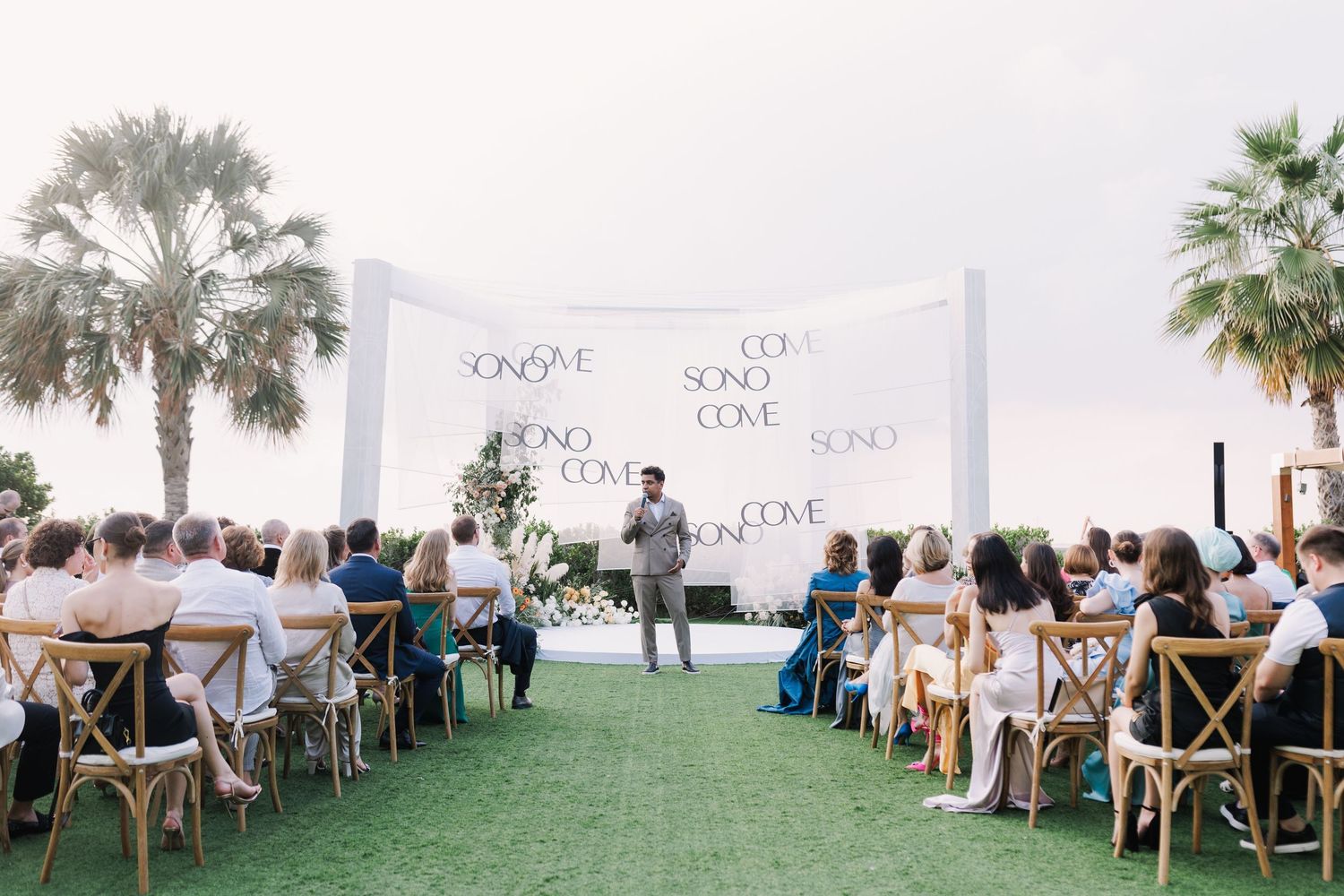 Wedding ceremony setup with white chairs and floral arrangements on a green lawn under palm trees.