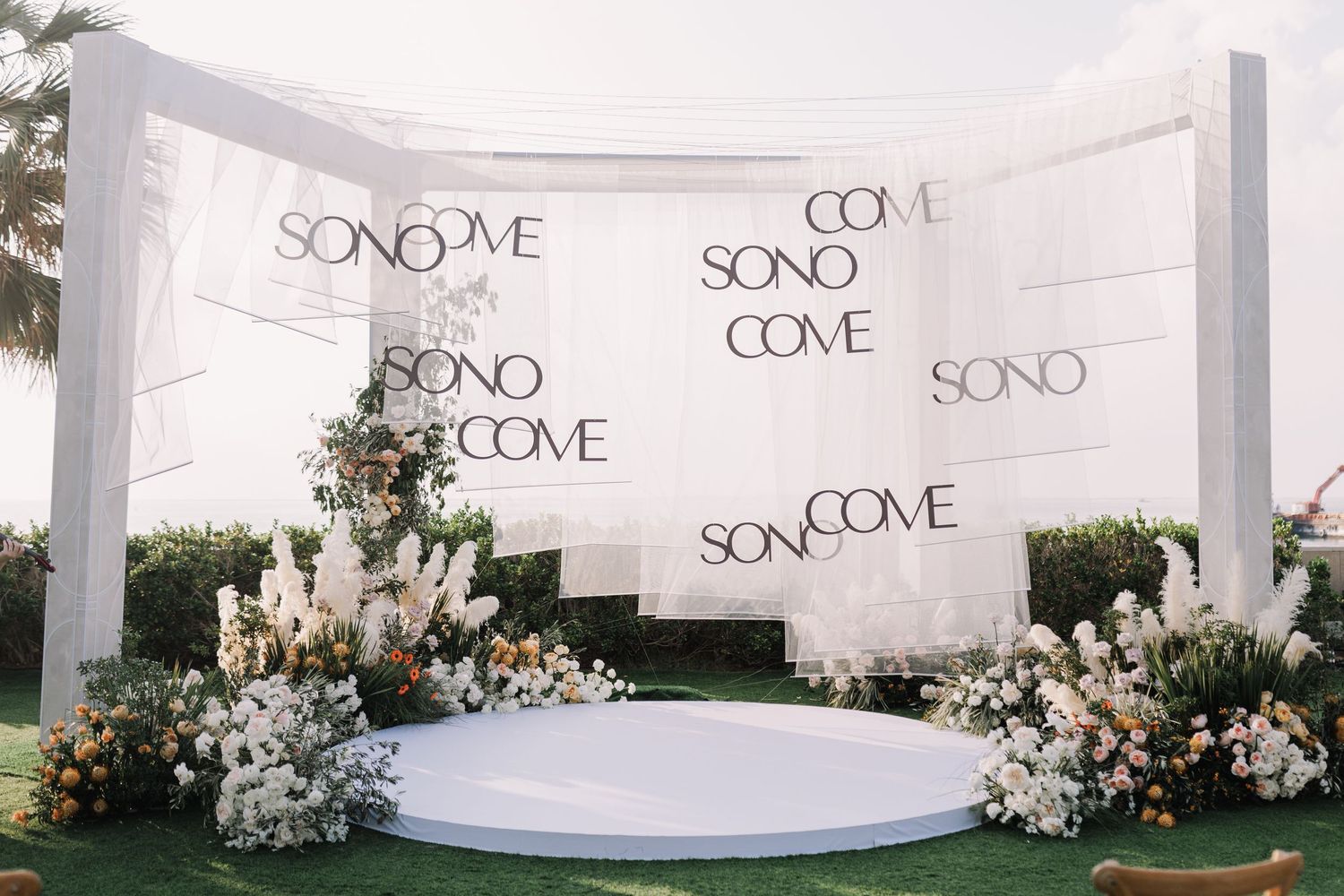 White curved wedding backdrop with floral arrangements and text display.