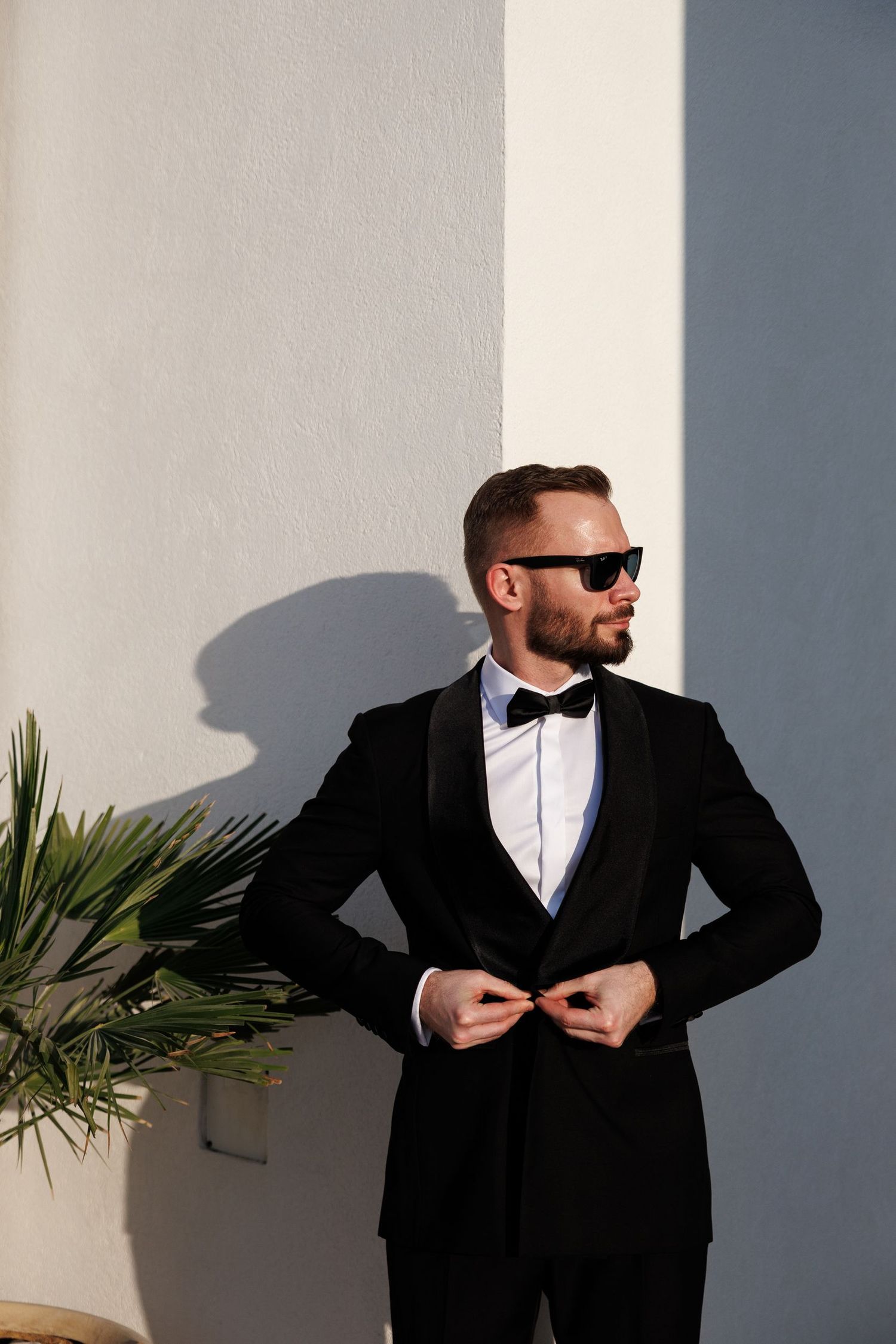 A groom in black tuxedo poses dramatically against a white wall creating strong shadows.