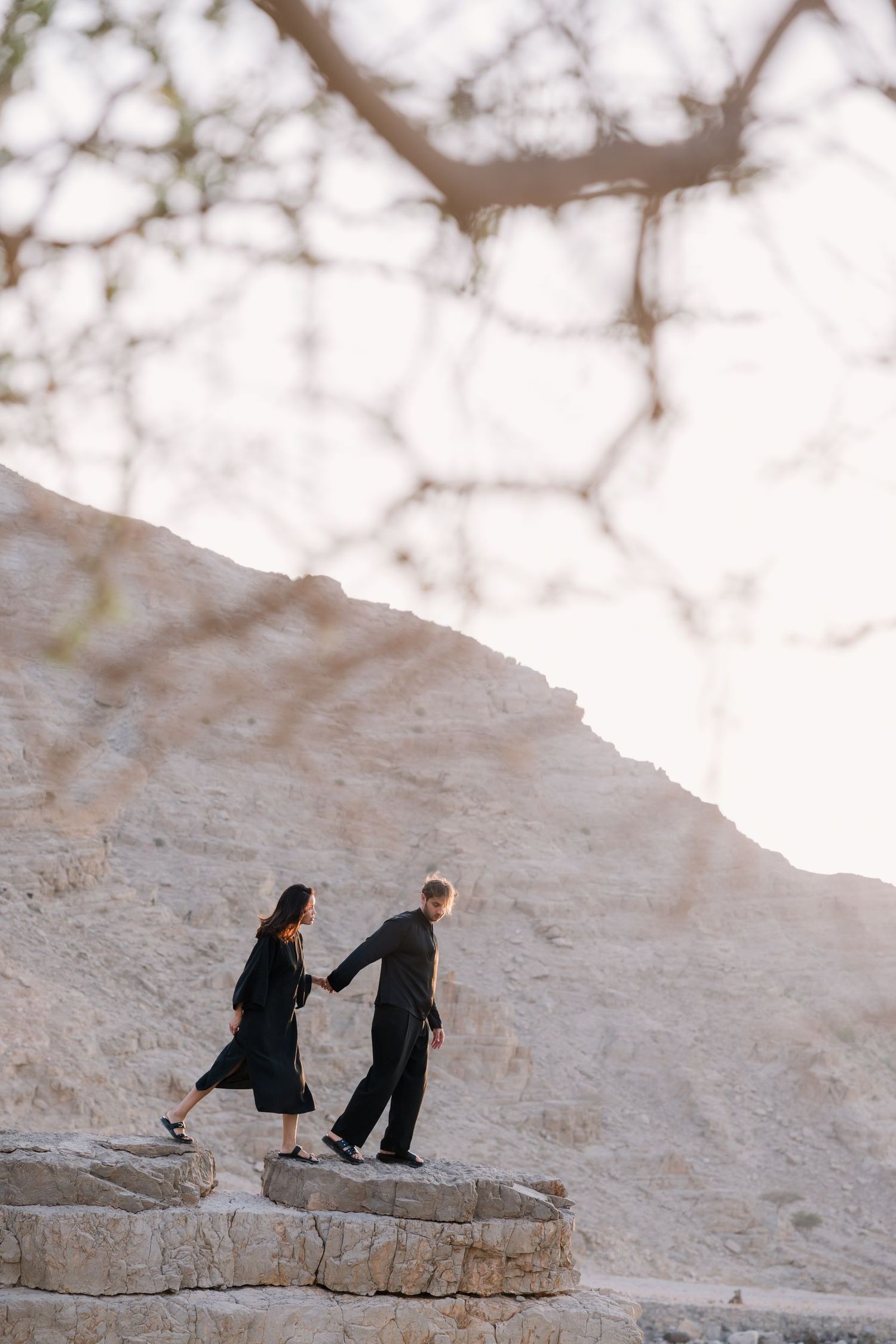 A couple walking hand in hand on rocky terrain against a bright desert landscape.