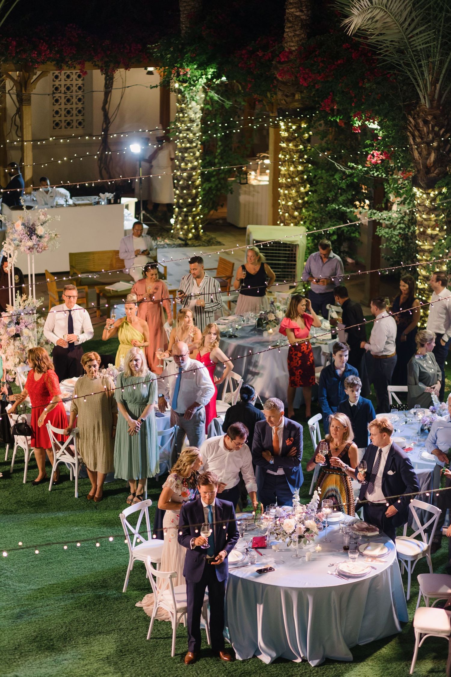 Evening wedding reception with guests dining under string lights and floral decorations.