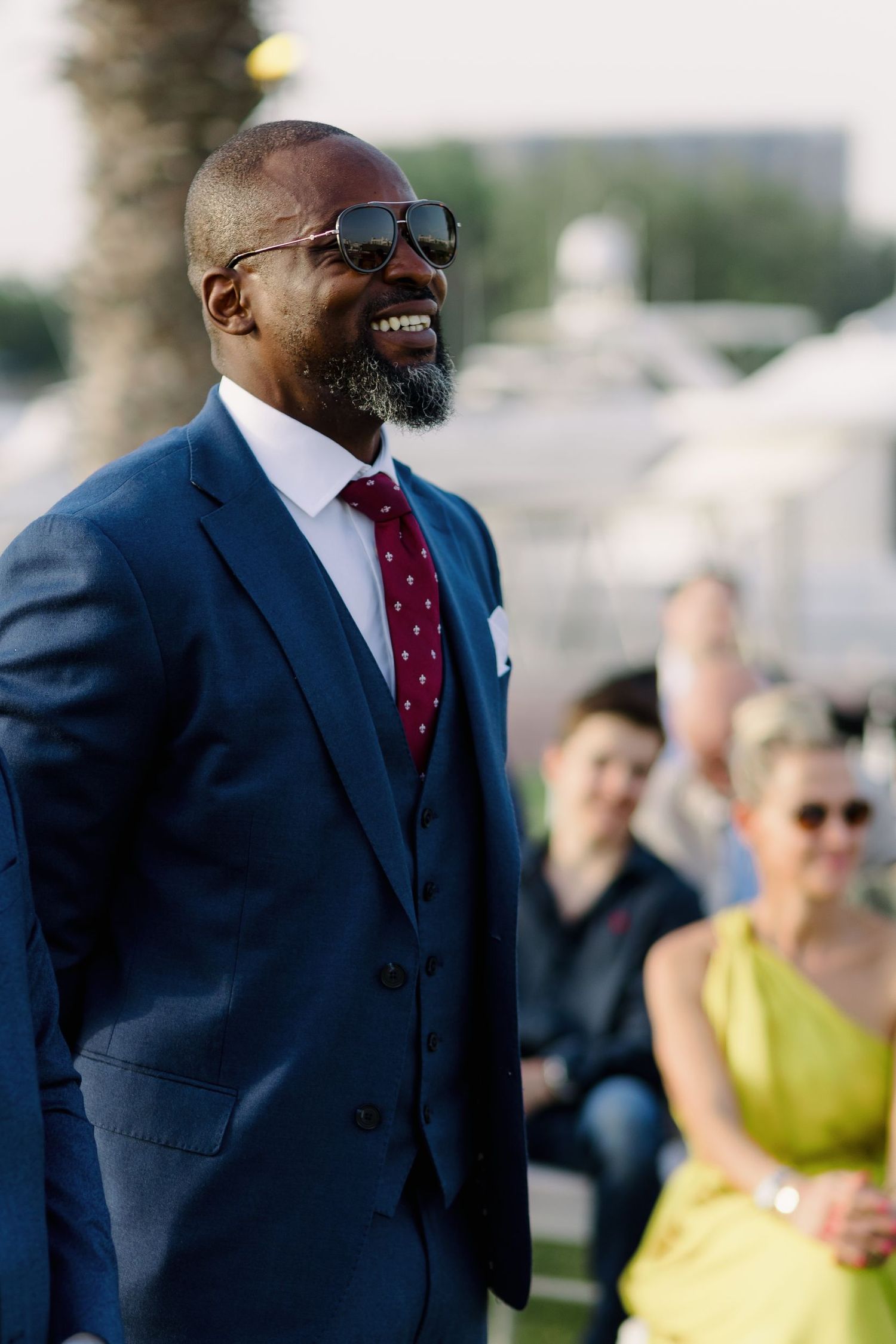 A guest in a navy suit and red tie smiling at an outdoor wedding celebration.