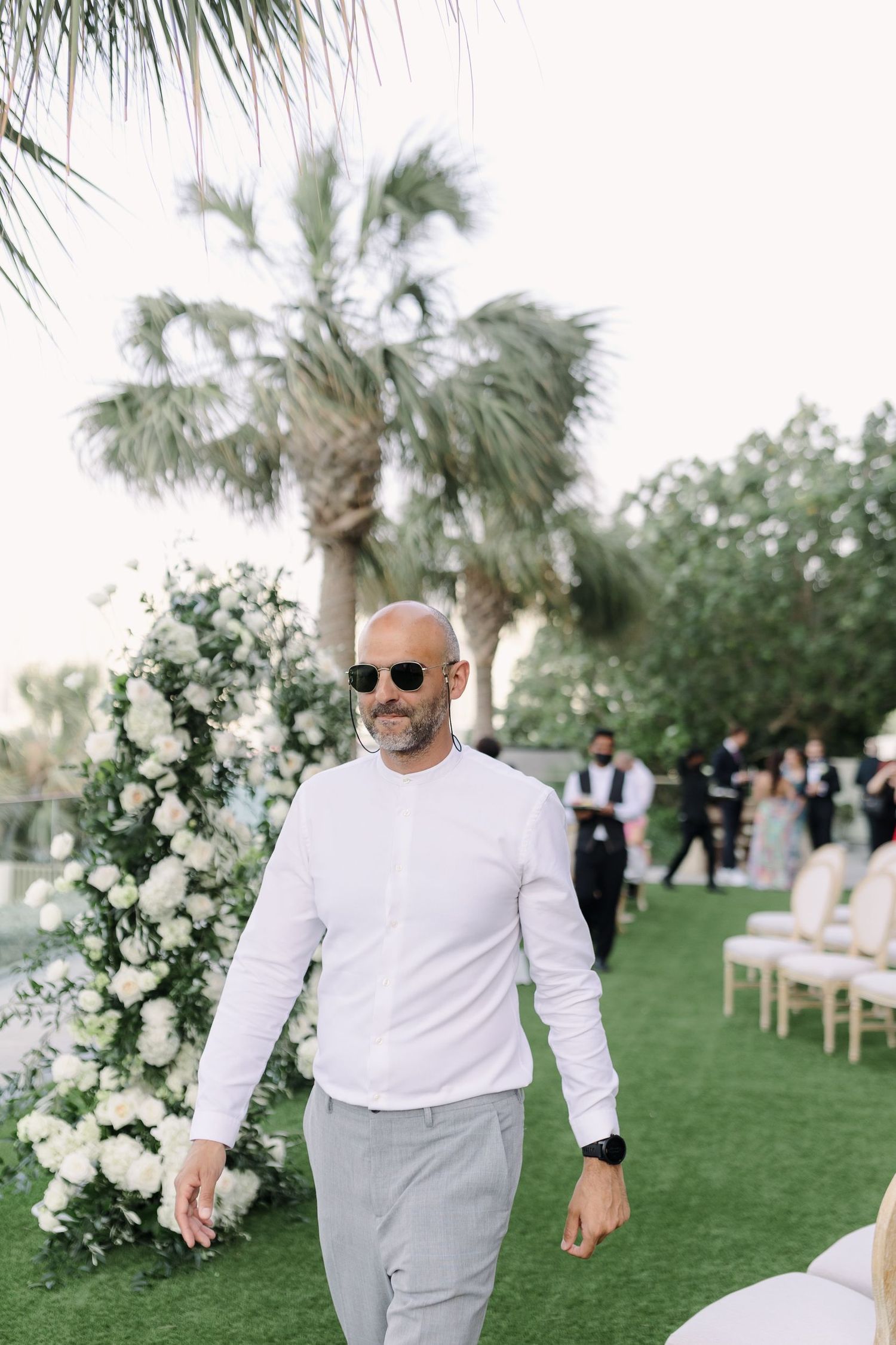 Wedding setup on a lawn with white floral arrangements and palm trees.
