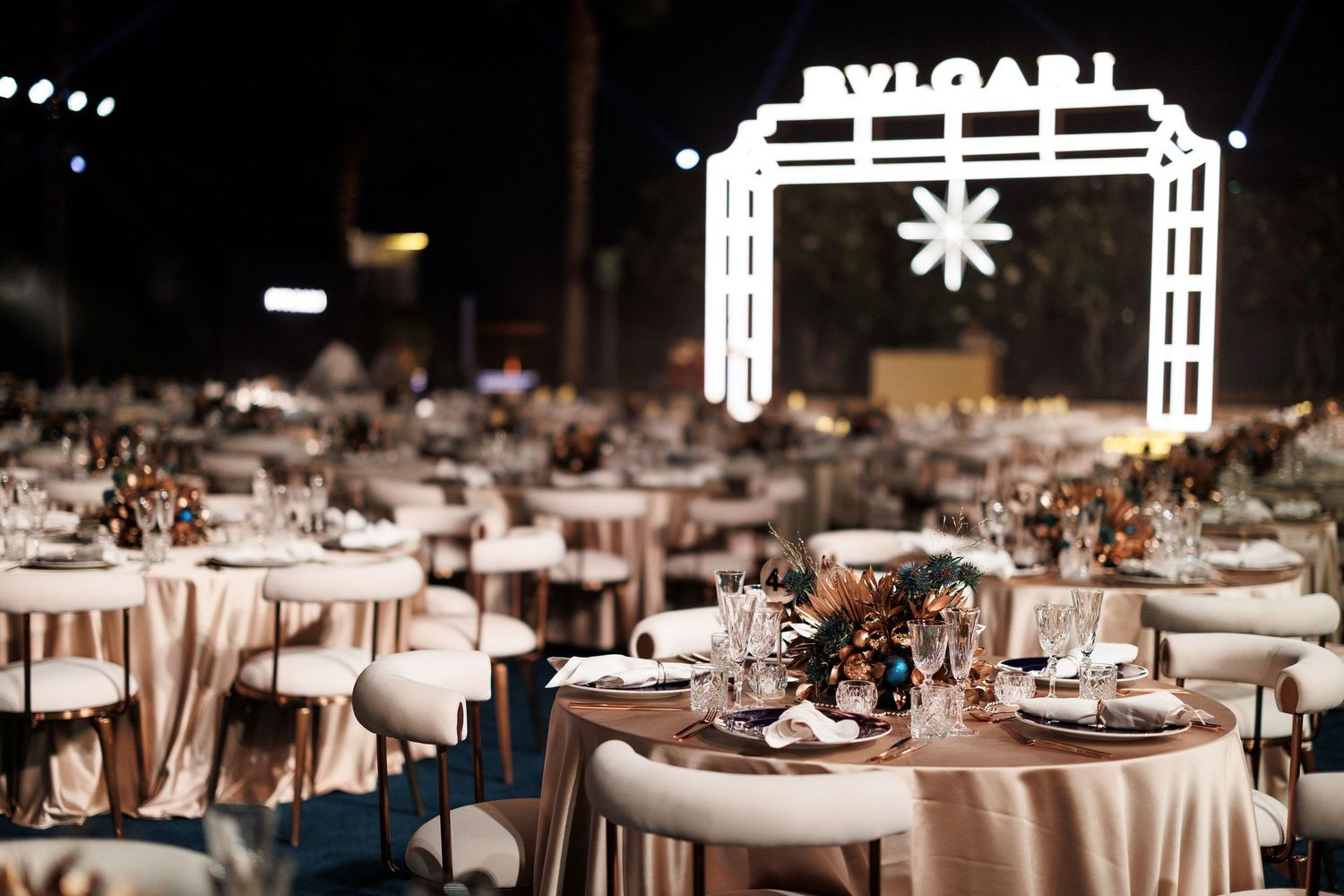 Elegant wedding reception setup with round tables and a glowing BVLGARI archway entrance at night.