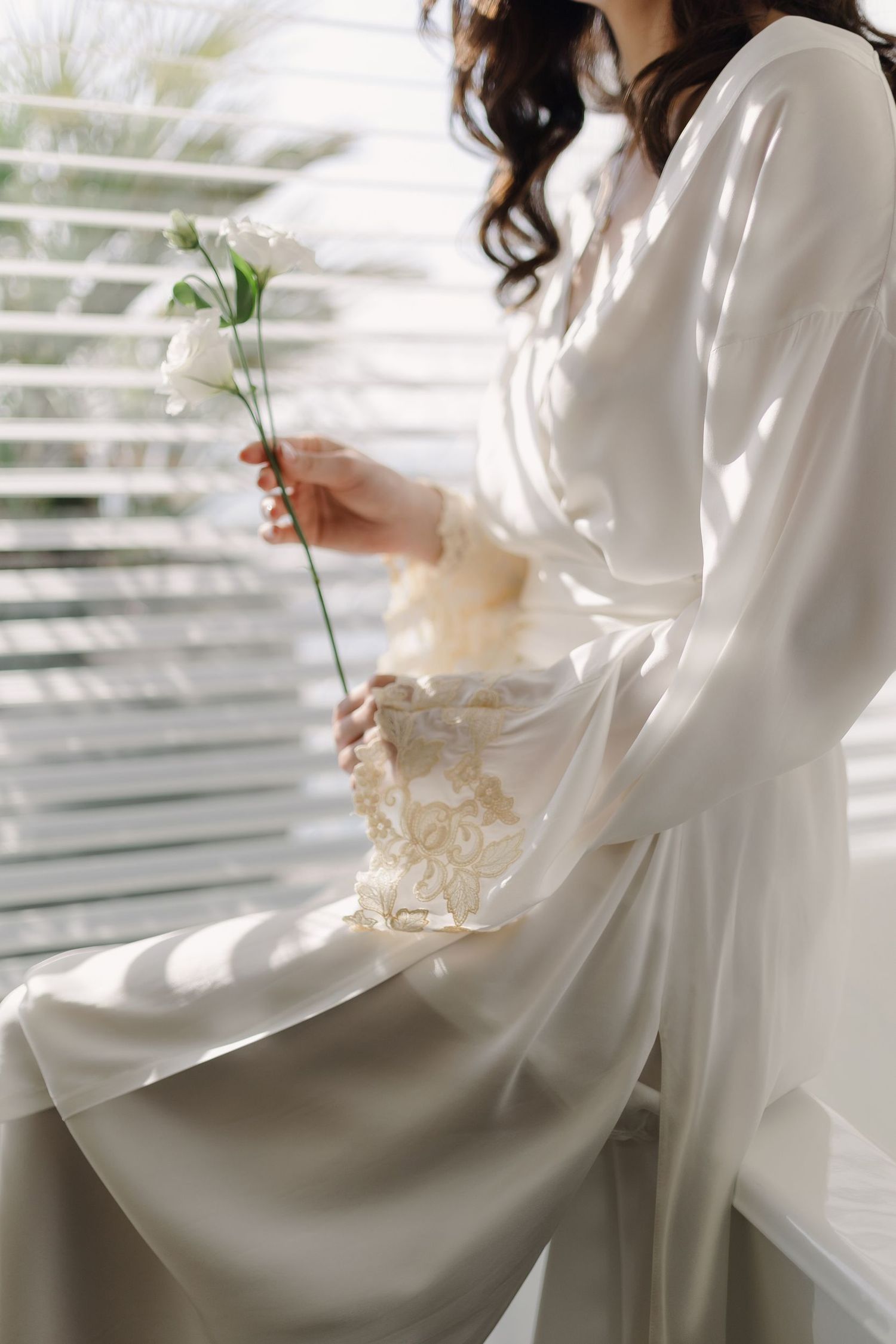 Figure in white clothing holding delicate flowers by a sunlit window with venetian blinds.