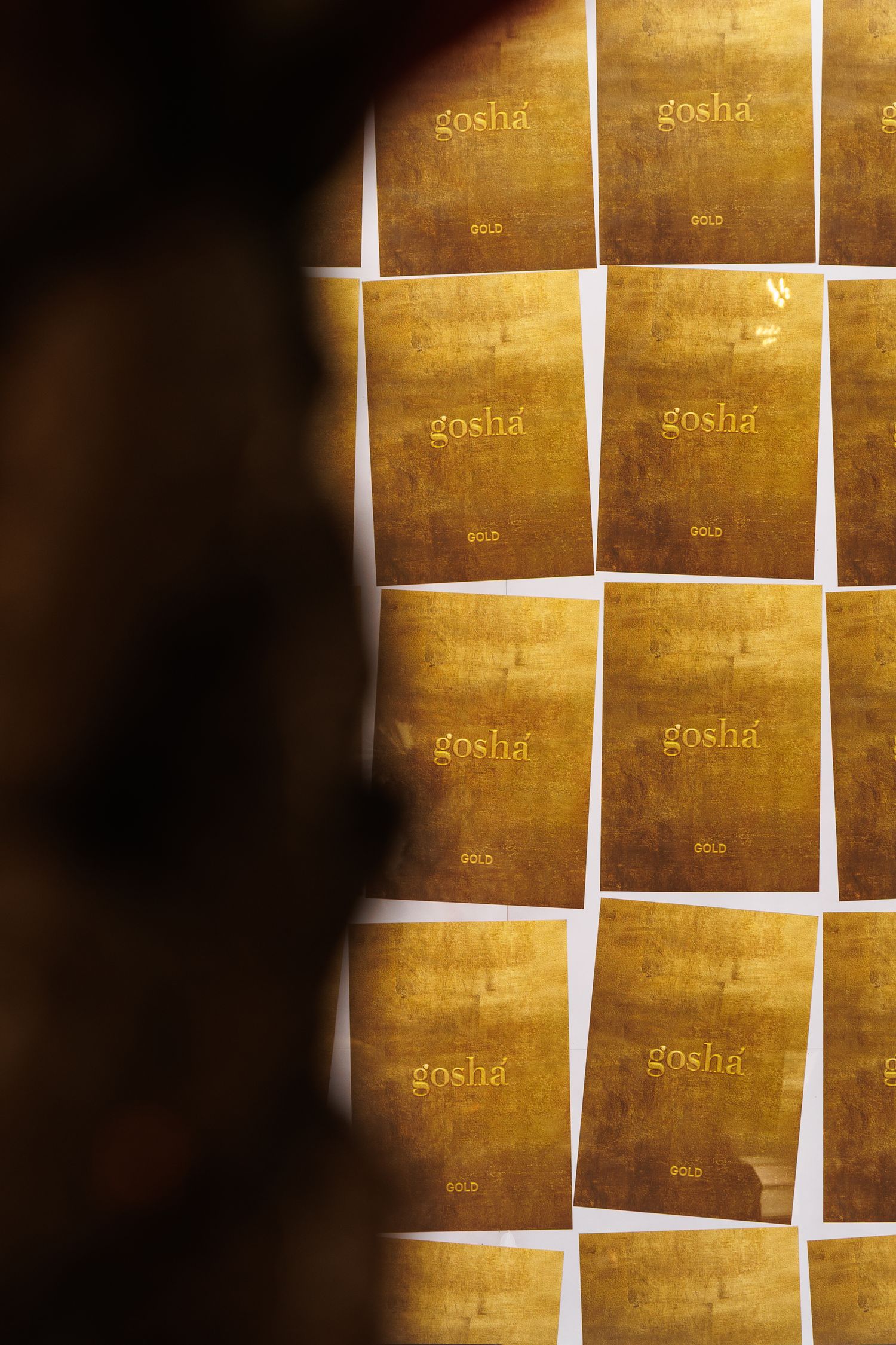 Grid of golden-tinted square prints with 'gosha' text arranged in rows against a dark background.