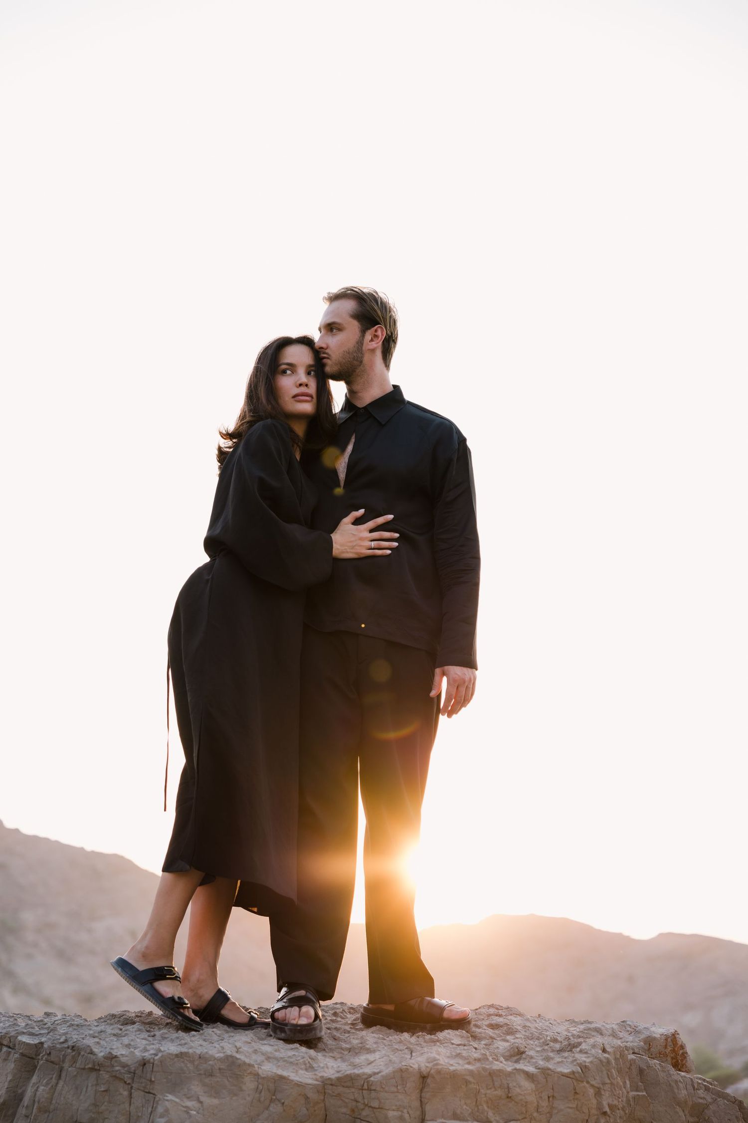 A couple embracing on a rocky outcrop with sunlight streaming behind them.