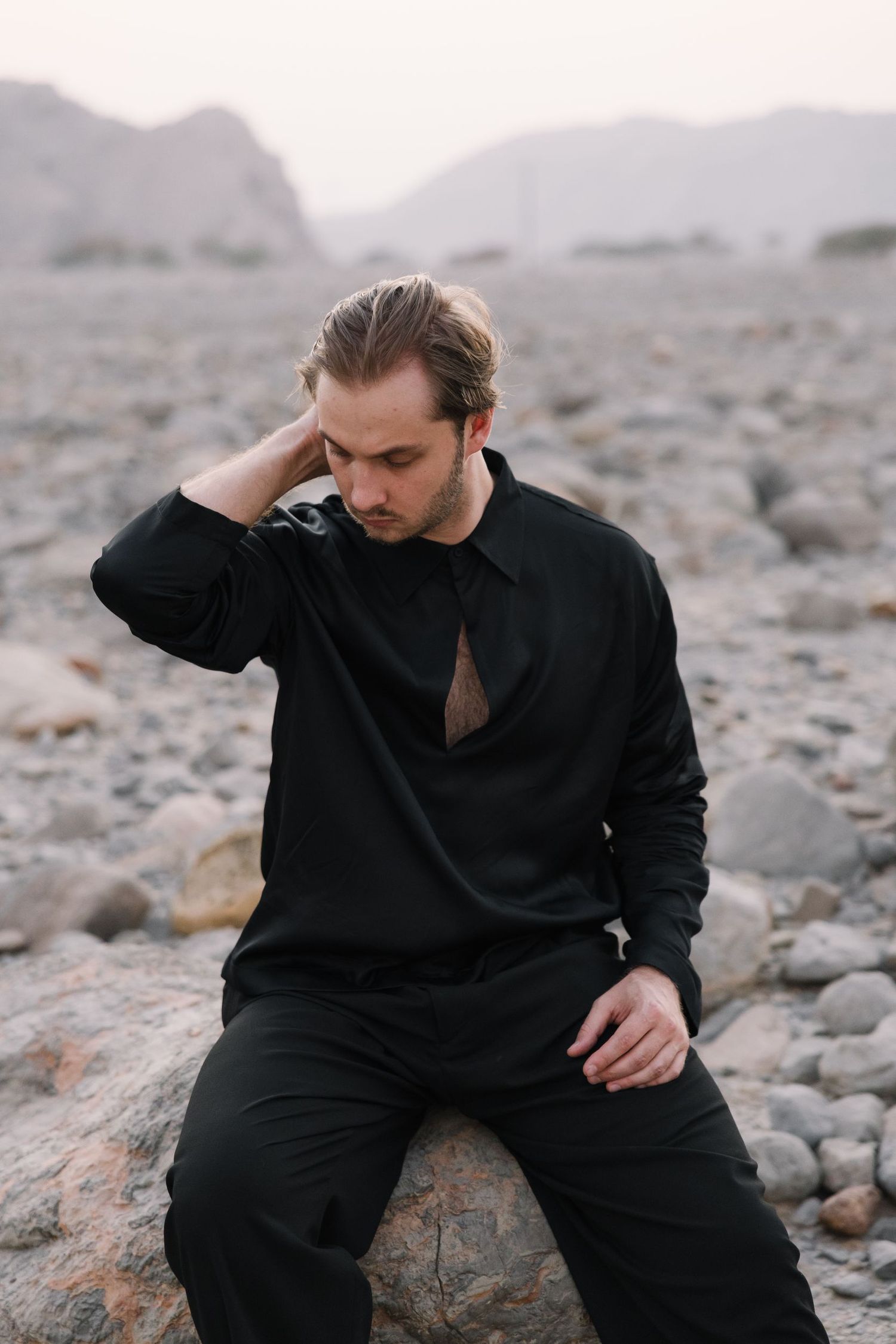 Figure in black clothing sits contemplatively on rocky terrain with mountains in background.