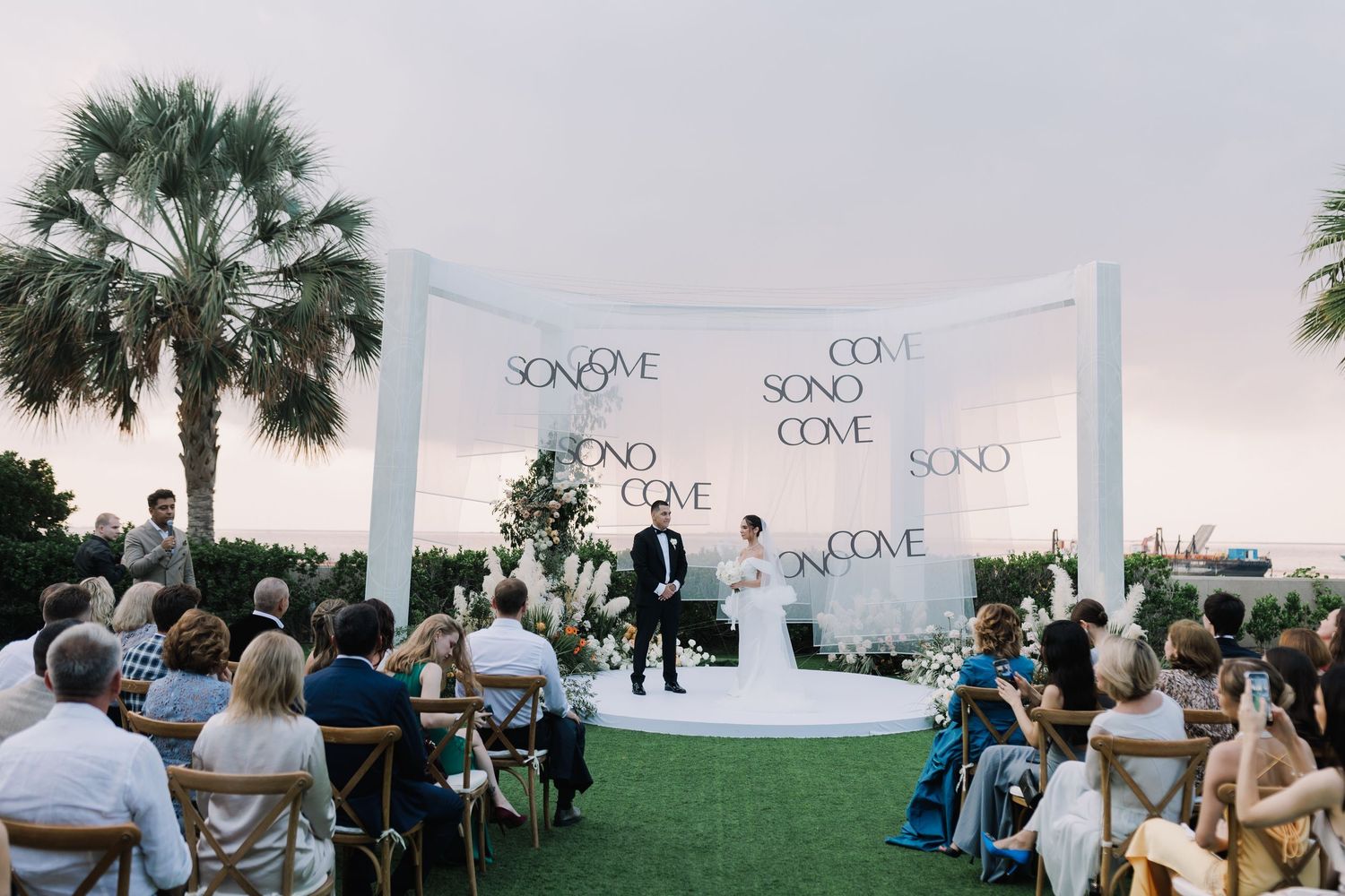 Panoramic view of an outdoor wedding ceremony with white backdrop and palm trees.