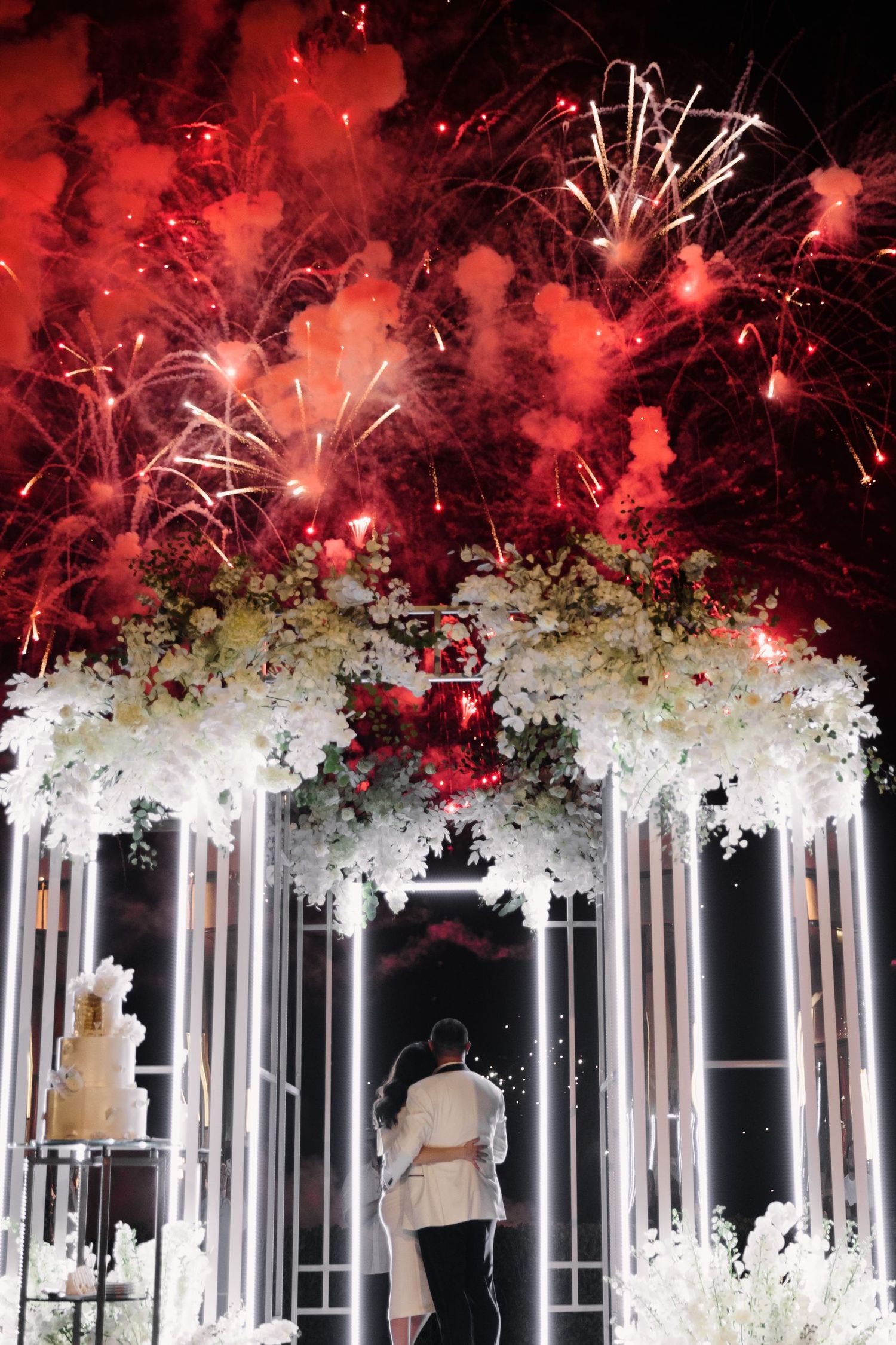 Elegant white floral archway with dramatic red fireworks bursting overhead.