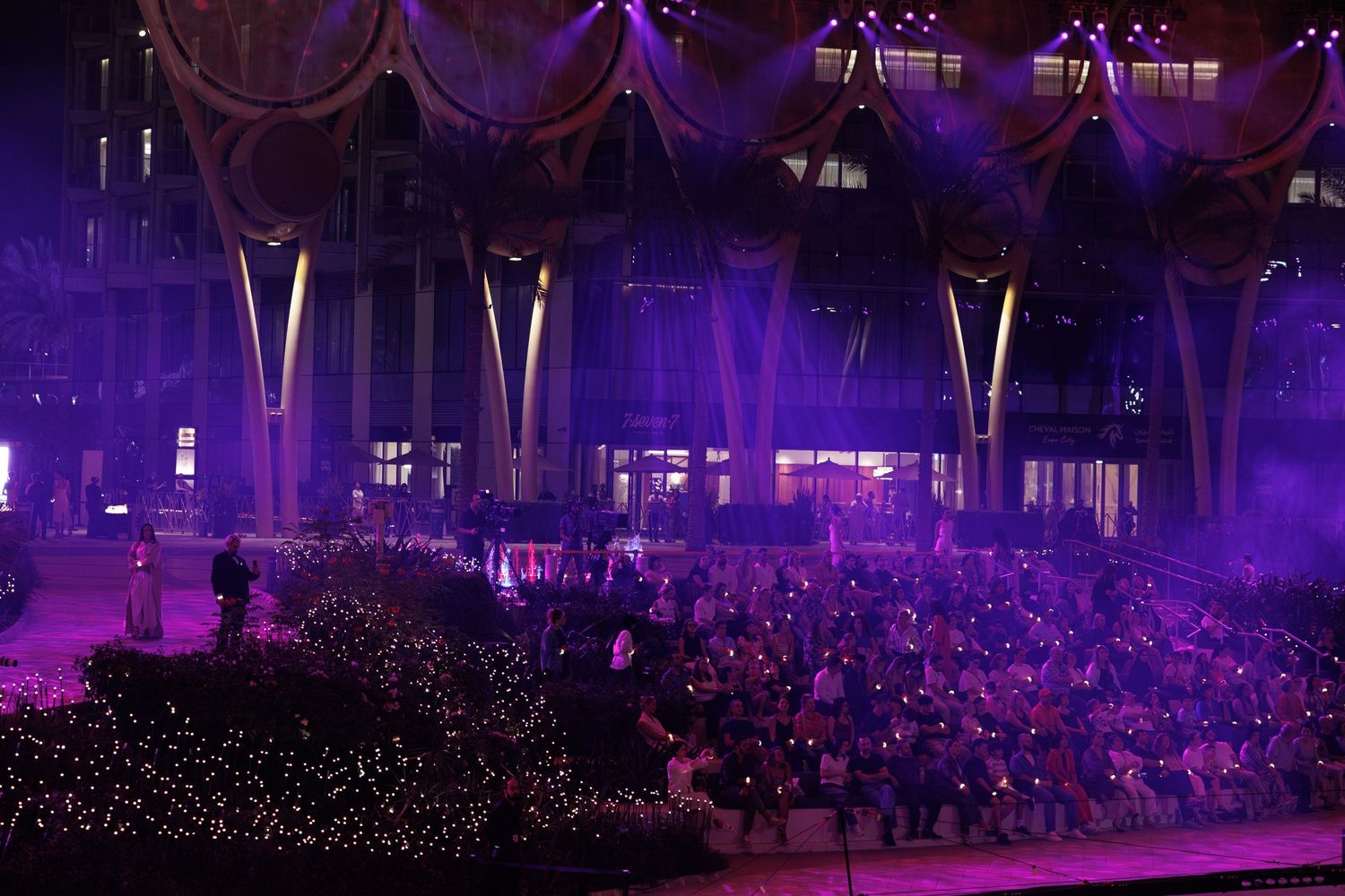 Concert venue illuminated in purple lighting with audience members visible in the background.