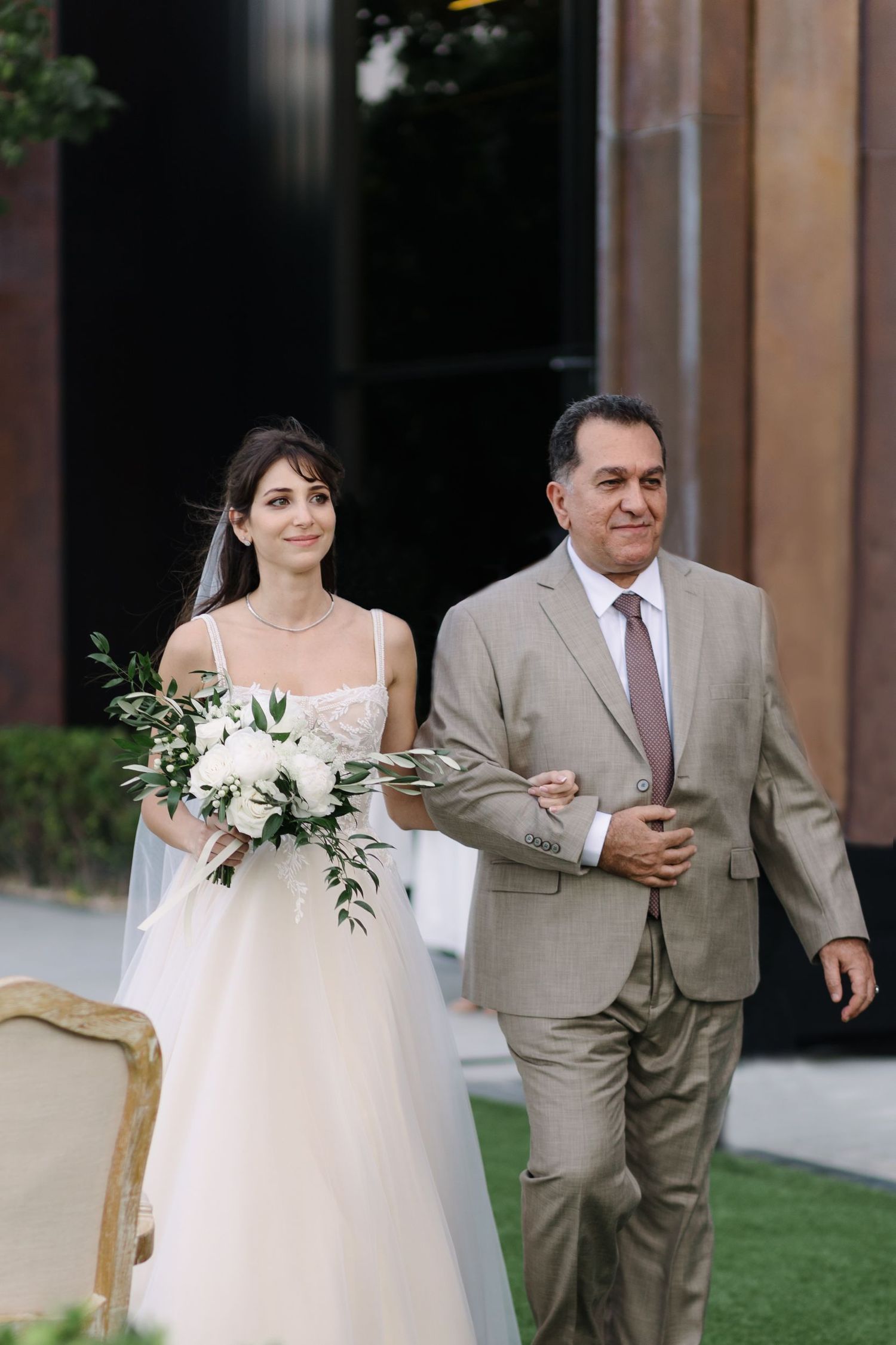 A bride in a flowing white gown walks with attendants in light beige suits.