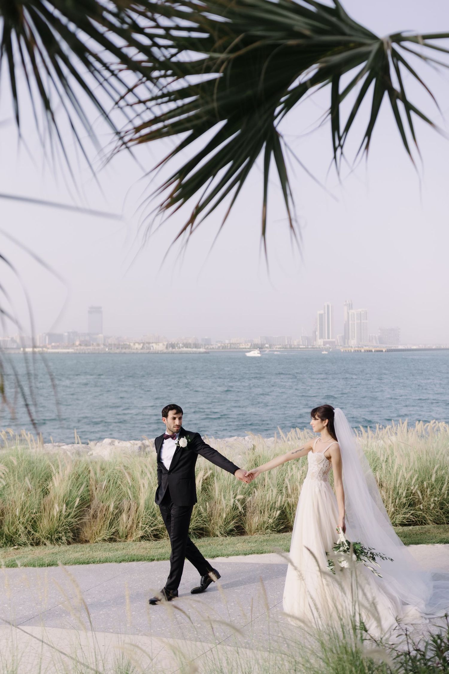 A romantic wedding couple poses by the waterfront with palm trees and city skyline in the background.