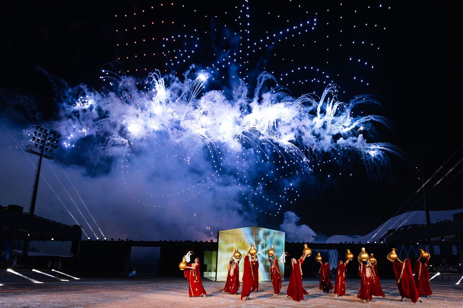 Spectacular fireworks display illuminates the night sky over a stadium with performers in red costumes on the field.