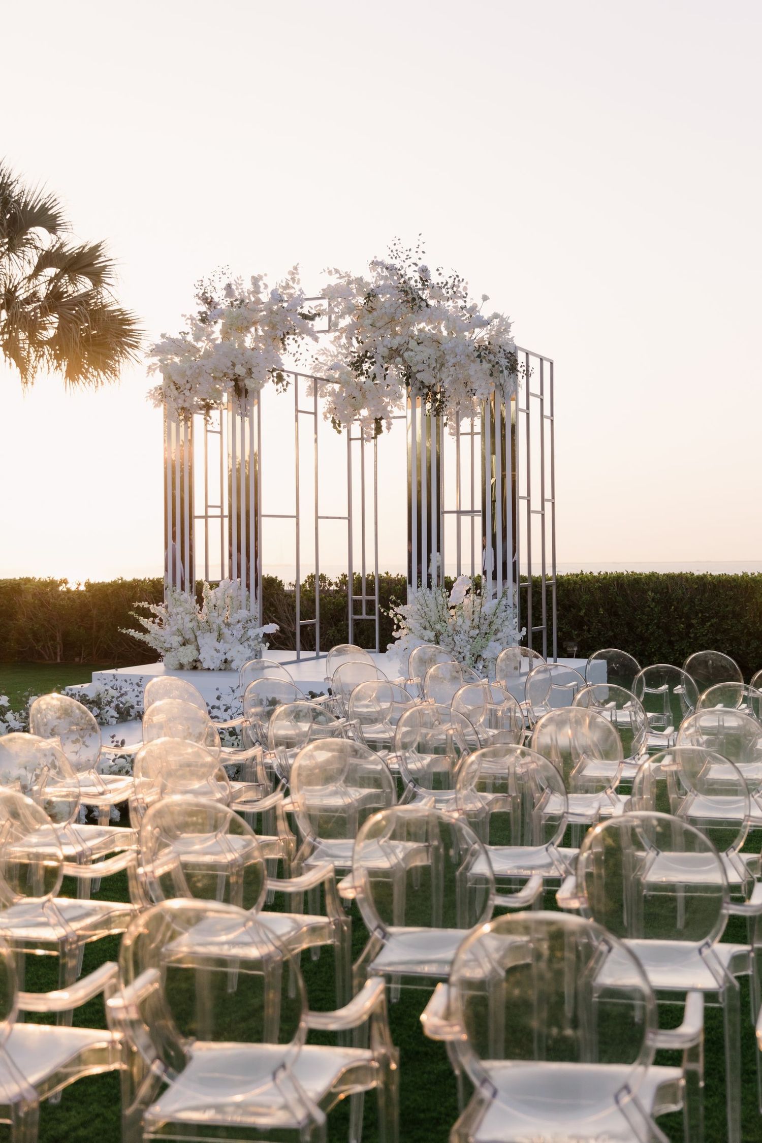Modern white wedding ceremony setup with clear chairs and floral arch against ocean backdrop.