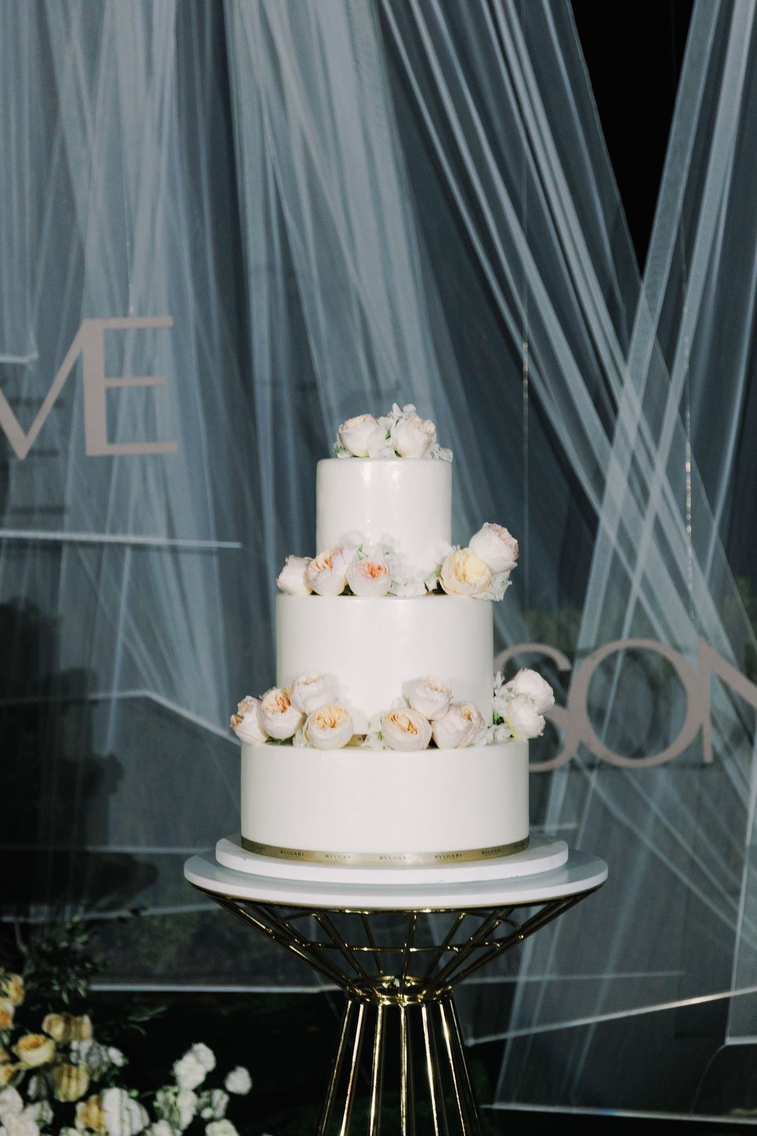 Elegant white wedding cake with fresh roses displayed against a light blue tulle backdrop.