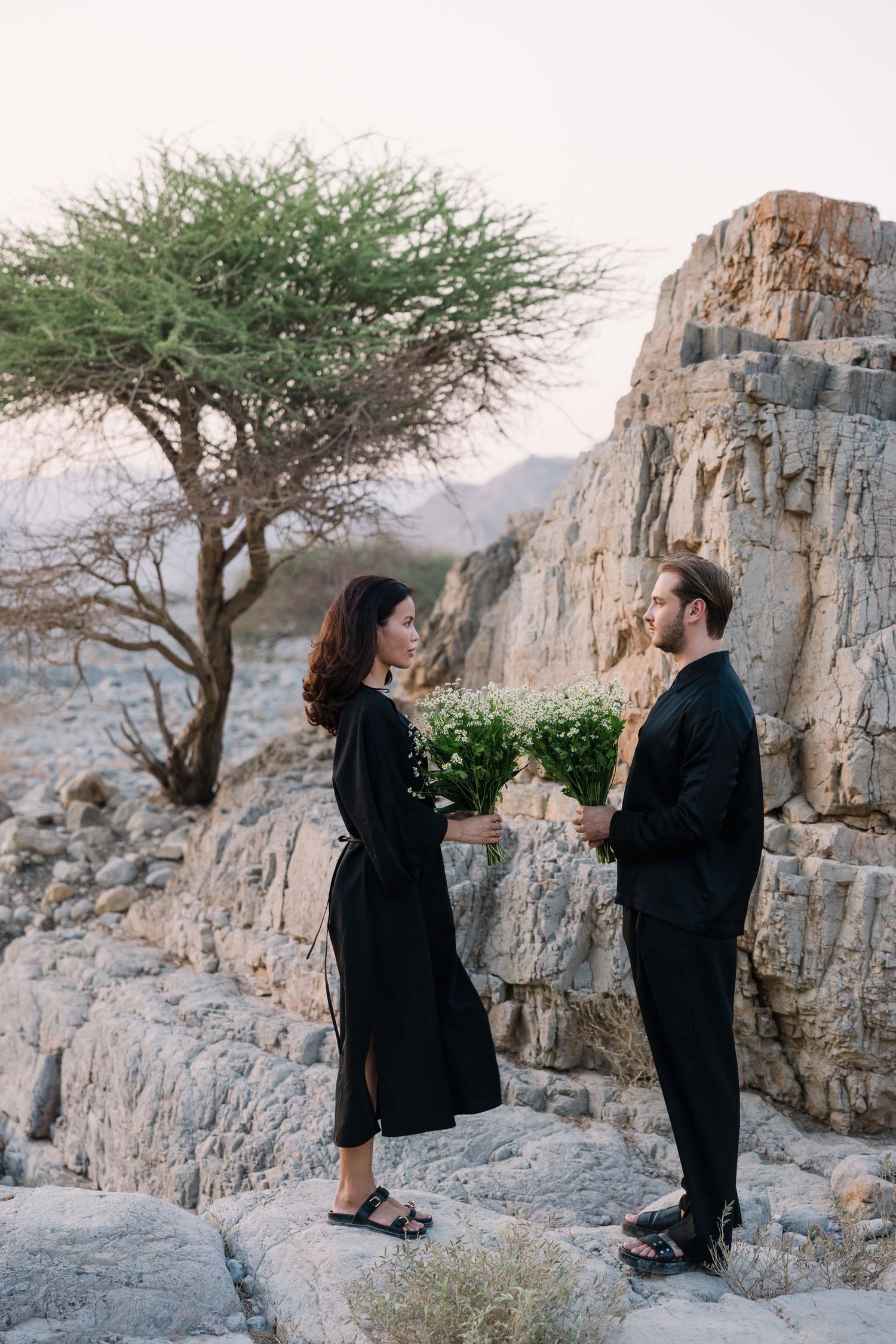 A couple standing on rocky terrain with a lone tree and mountain backdrop.