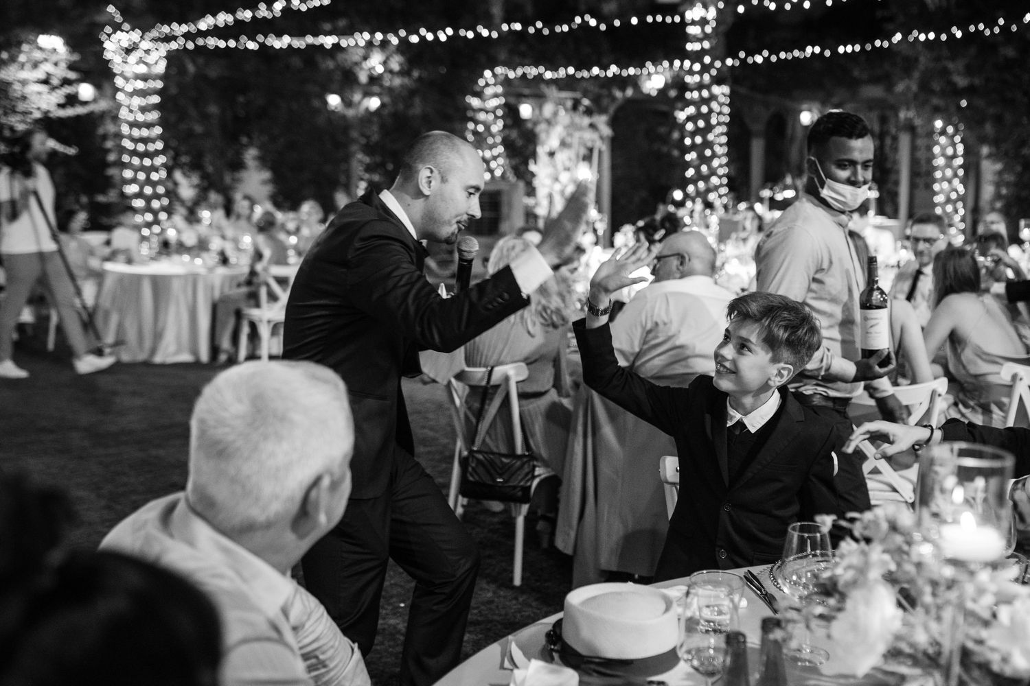 Black and white photo of wedding guests enjoying an evening reception under string lights.