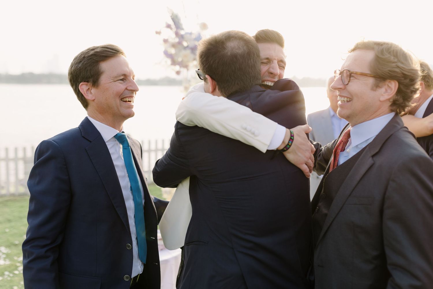 A joyful group embrace during an outdoor wedding celebration by the water.