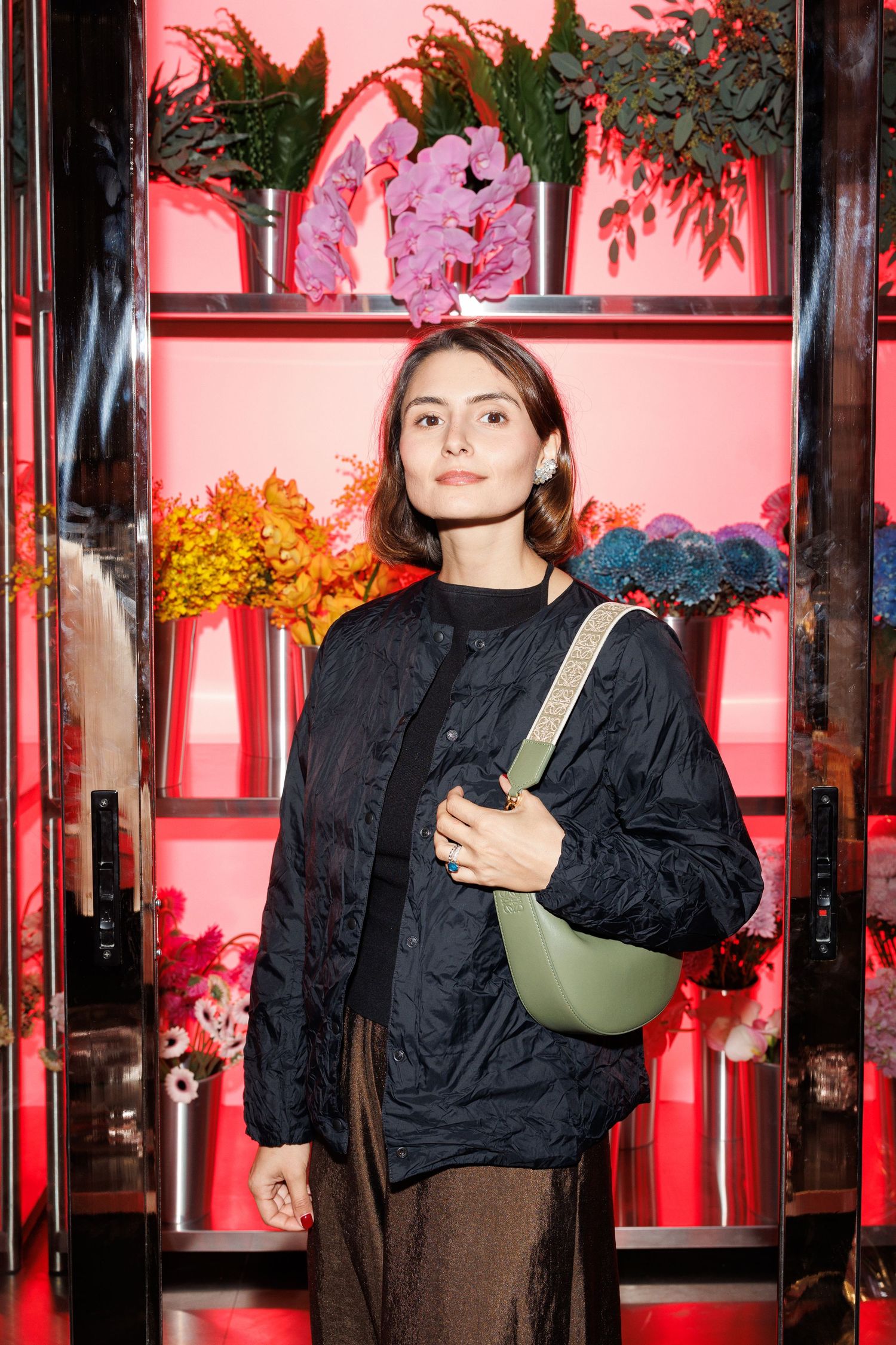 Person in black jacket poses against pink-lit backdrop with autumn foliage visible.