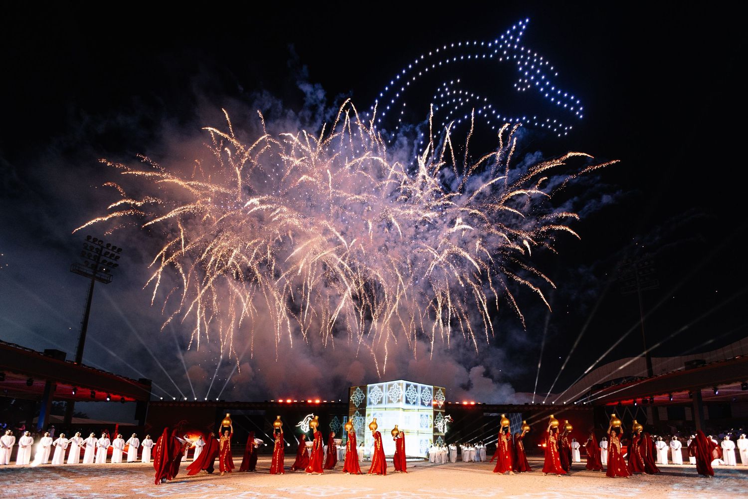 Fireworks display lighting up the night sky above a traditional performance with performers in white costumes.