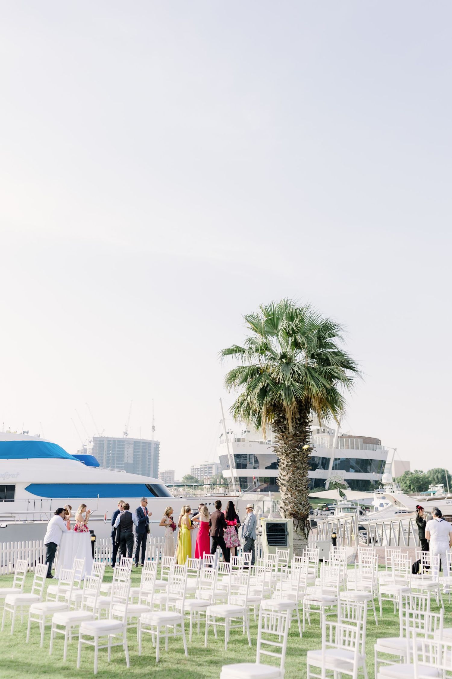 White chairs arranged for an outdoor wedding ceremony with palm trees and boats in the background.