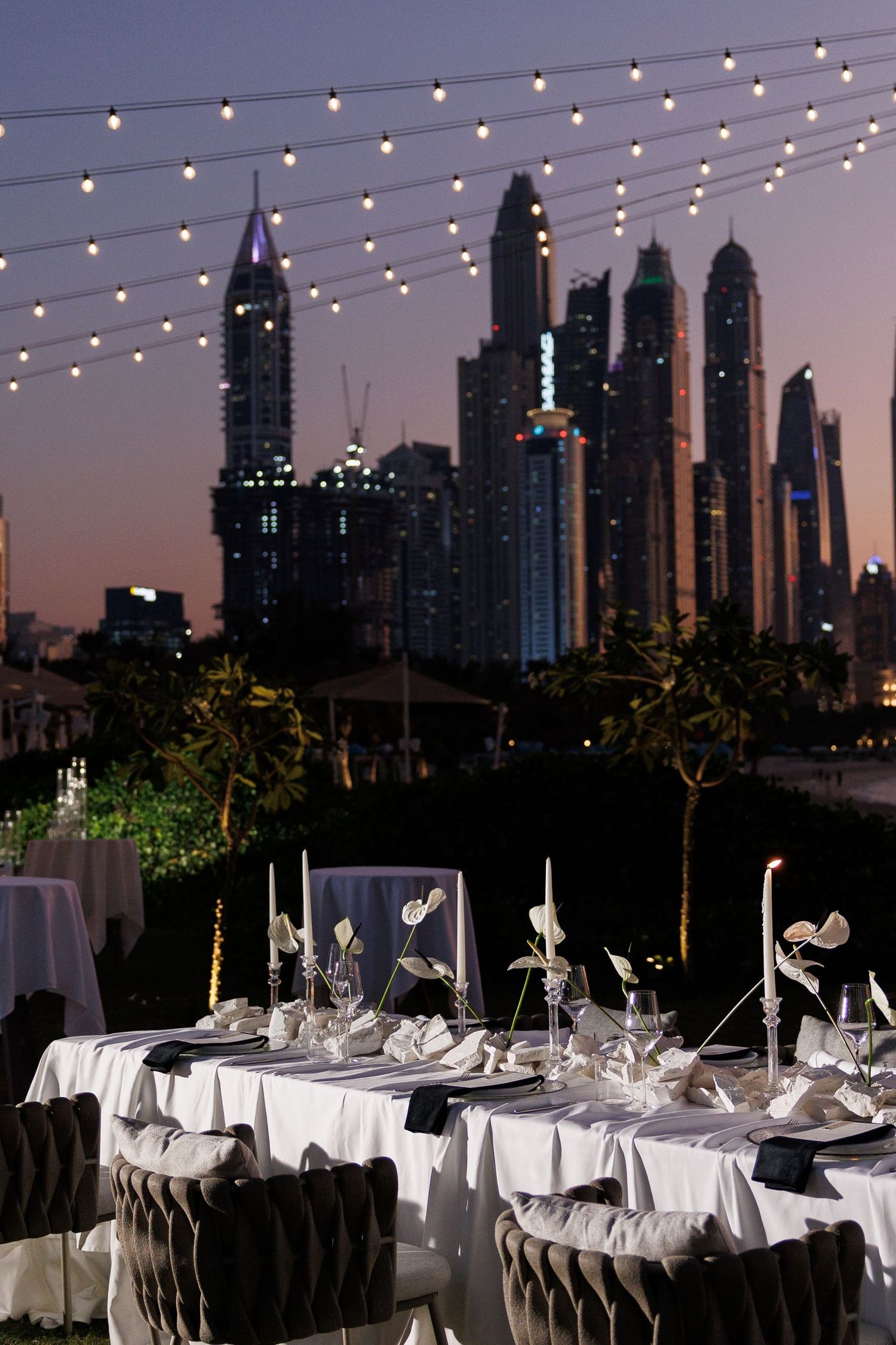 Elegant outdoor wedding reception setup with string lights and Dubai skyline at dusk.