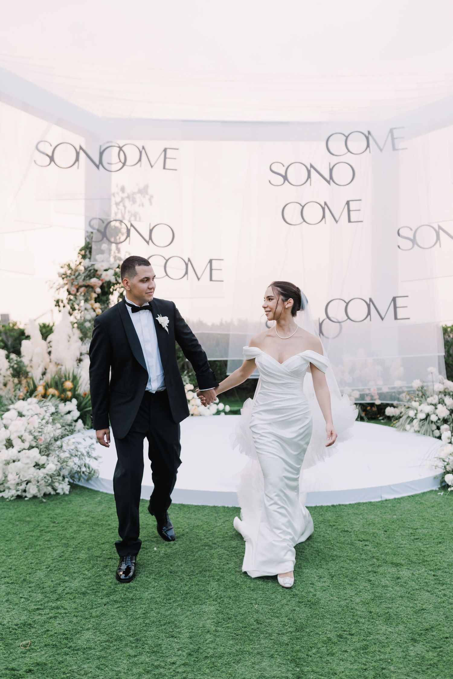 Newlyweds dancing on green lawn in front of white backdrop with 'SONO' text.