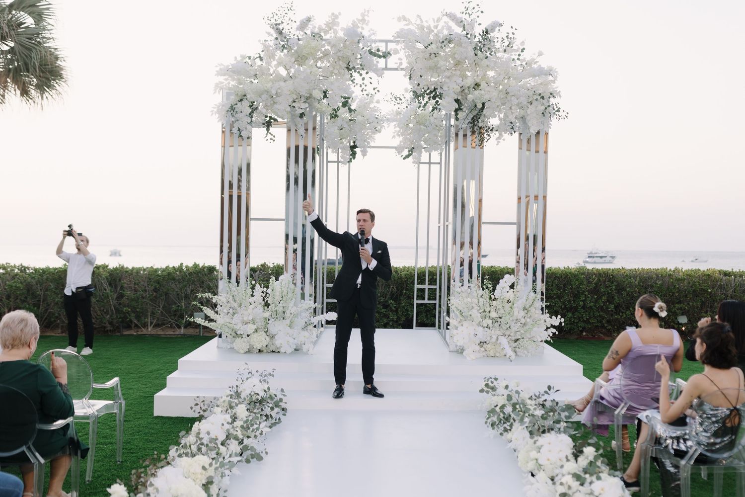Wedding ceremony with decorated white metal arch and clear chairs on oceanfront lawn.