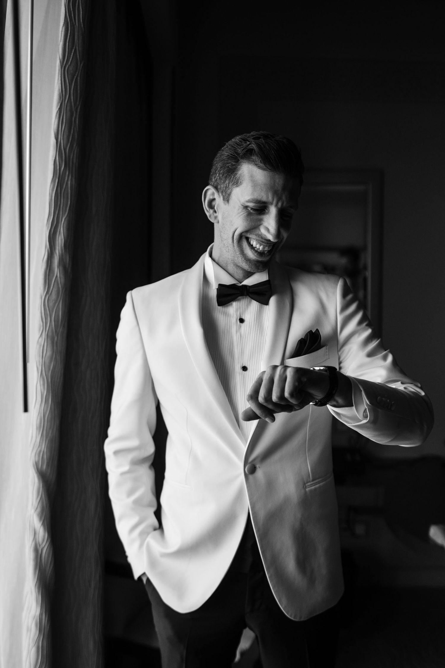 Groom in white tuxedo jacket and bowtie checks his watch while smiling near a window.