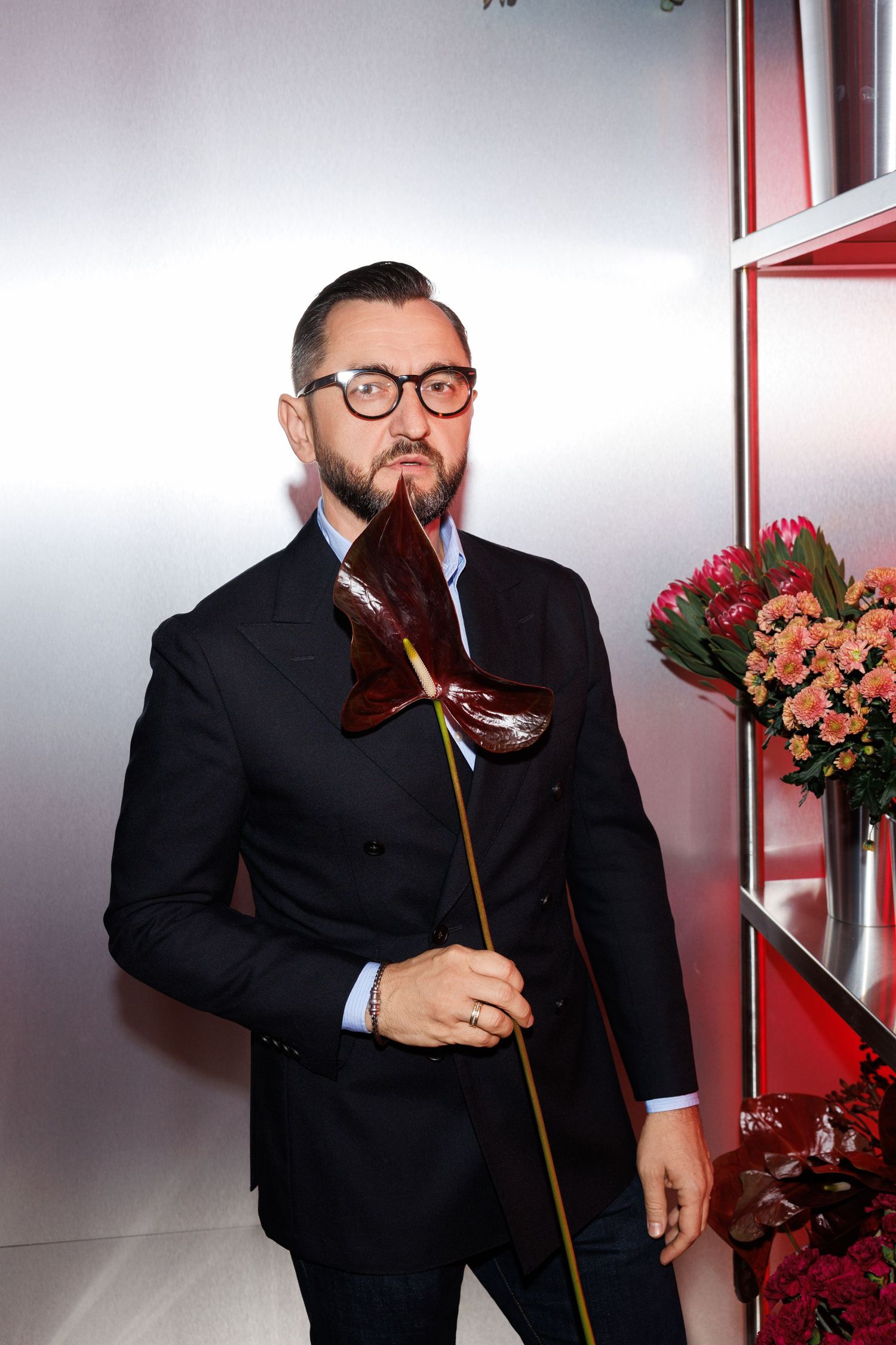 Person in formal black suit holding wine glass poses near floral arrangements.