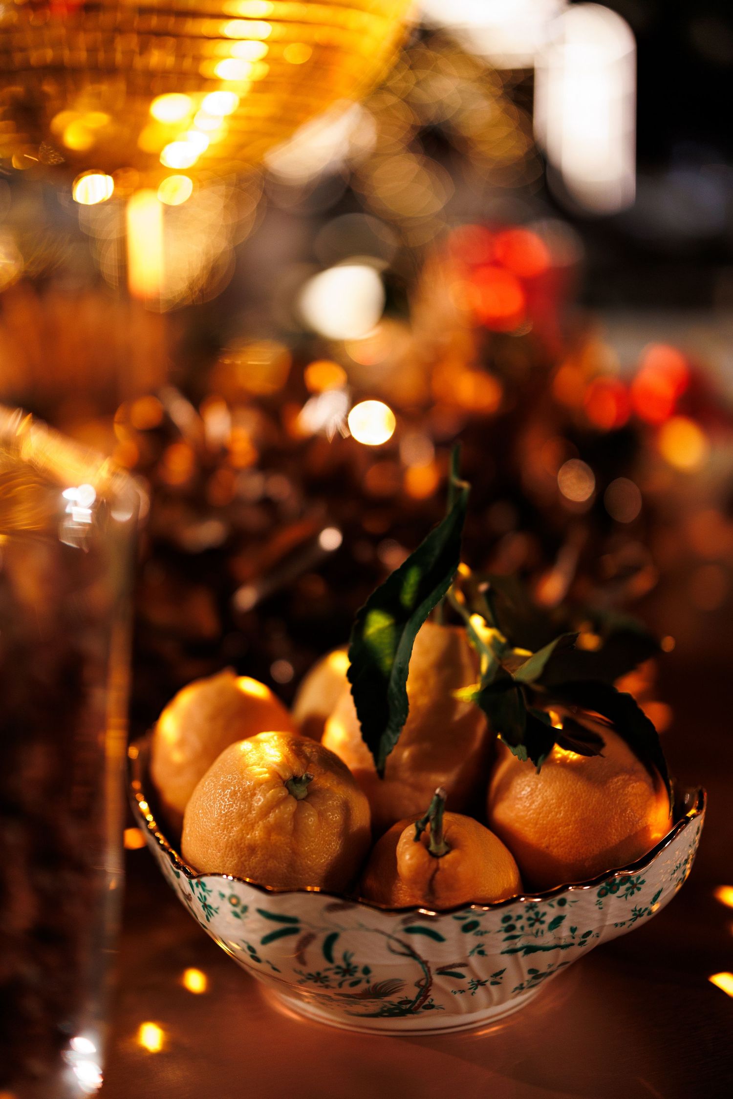 Fresh oranges with green leaves arranged in a decorative ceramic bowl with glowing holiday lights in the background.