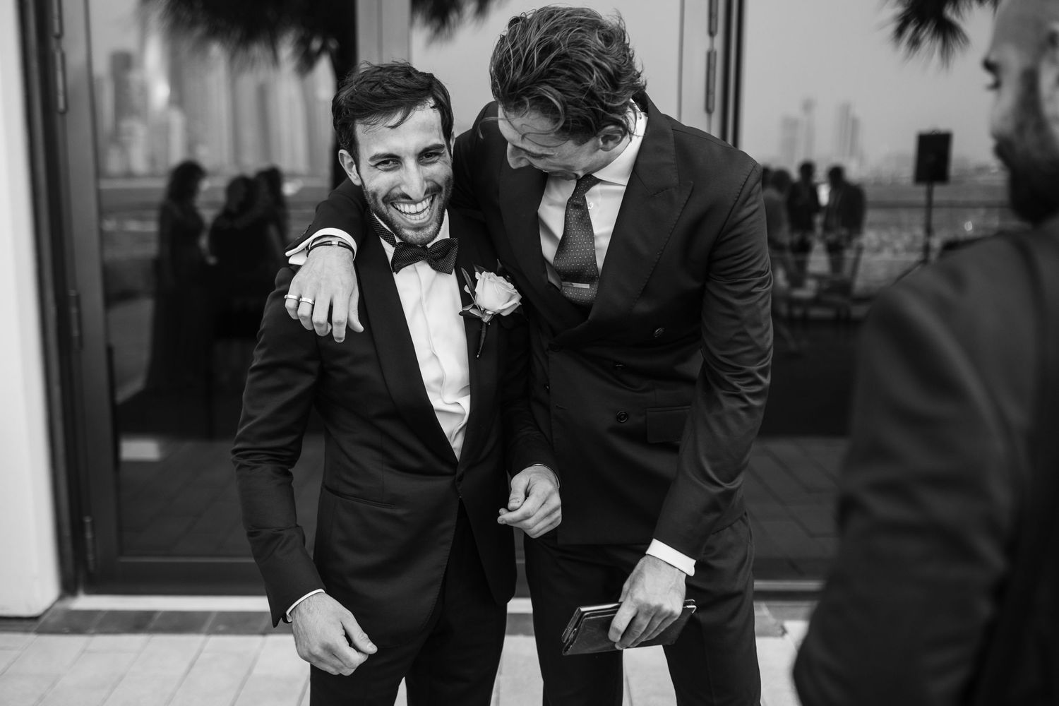 Groomsmen in black tuxedos share a candid laughing moment together at a wedding.