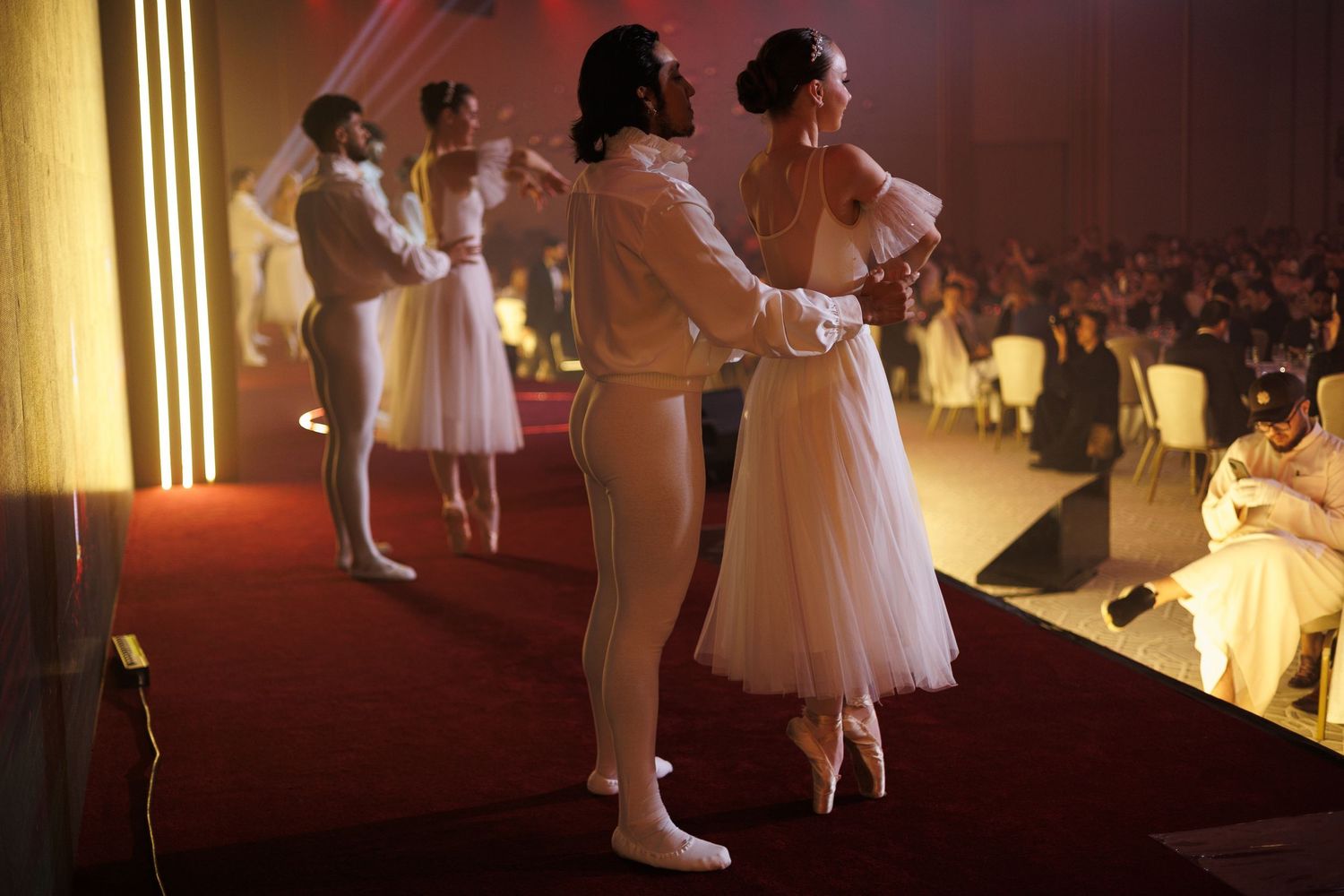 Ballet dancers in white costumes perform on illuminated stage with warm lighting.