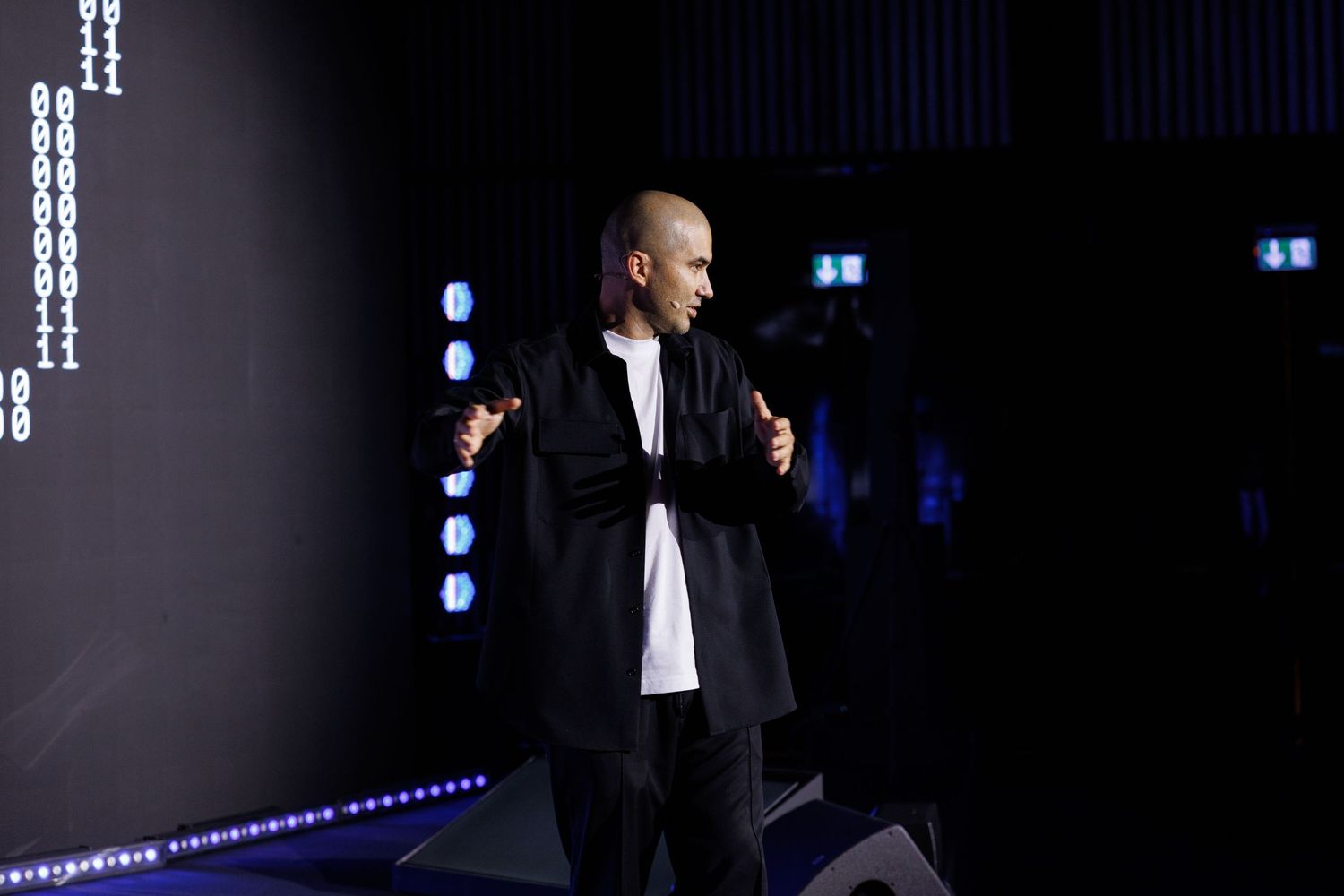 Performer speaks into microphone on darkened stage with dramatic backlighting.