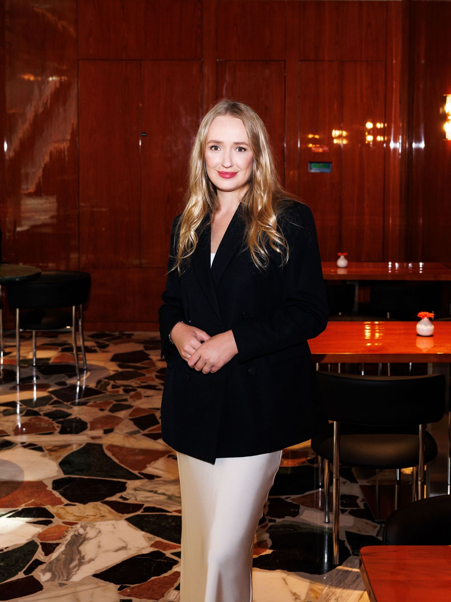 Person in black blazer poses against wood-paneled wall in restaurant setting.