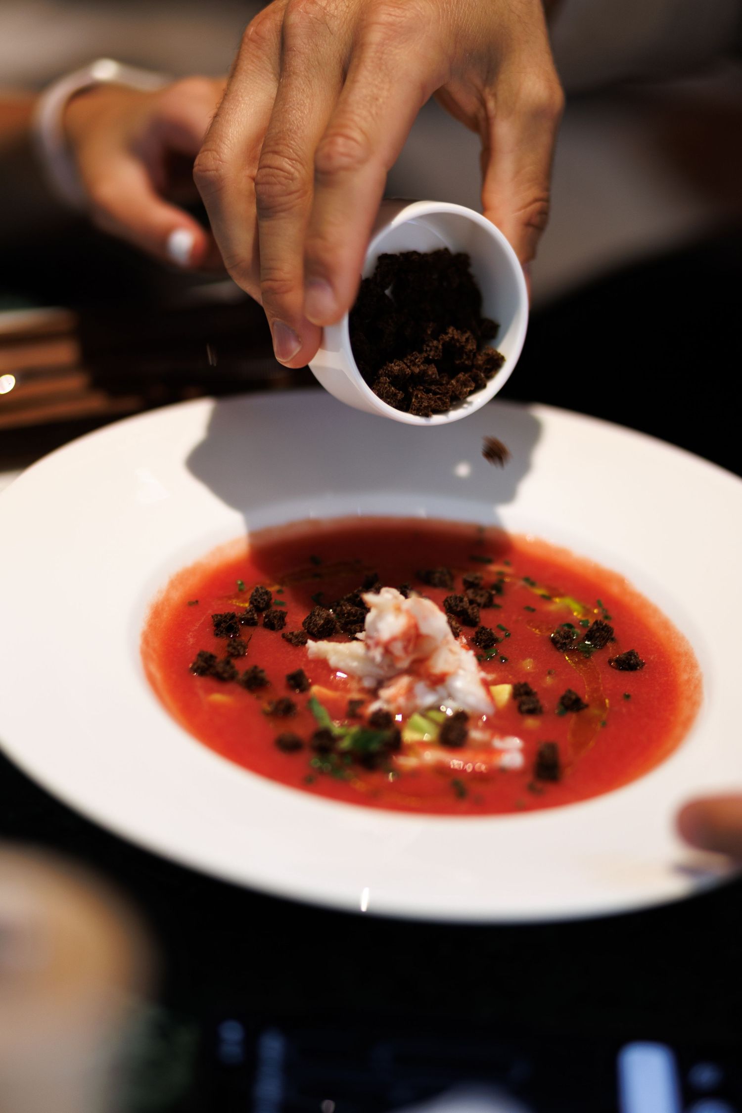 A hand garnishing a vibrant red soup with dark toppings in a white bowl.