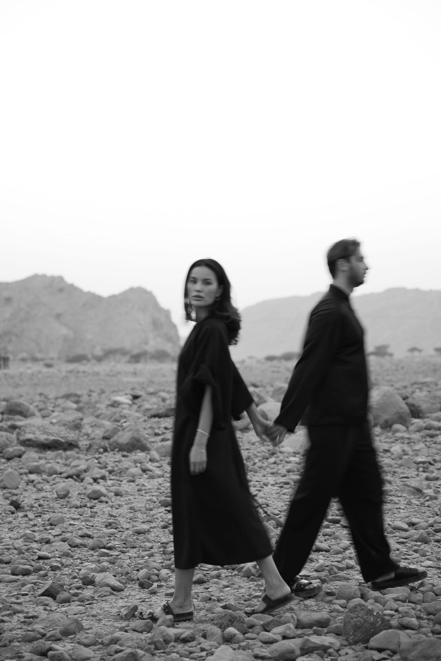 Figures in dark clothing walking across rocky desert terrain with mountains in distance.