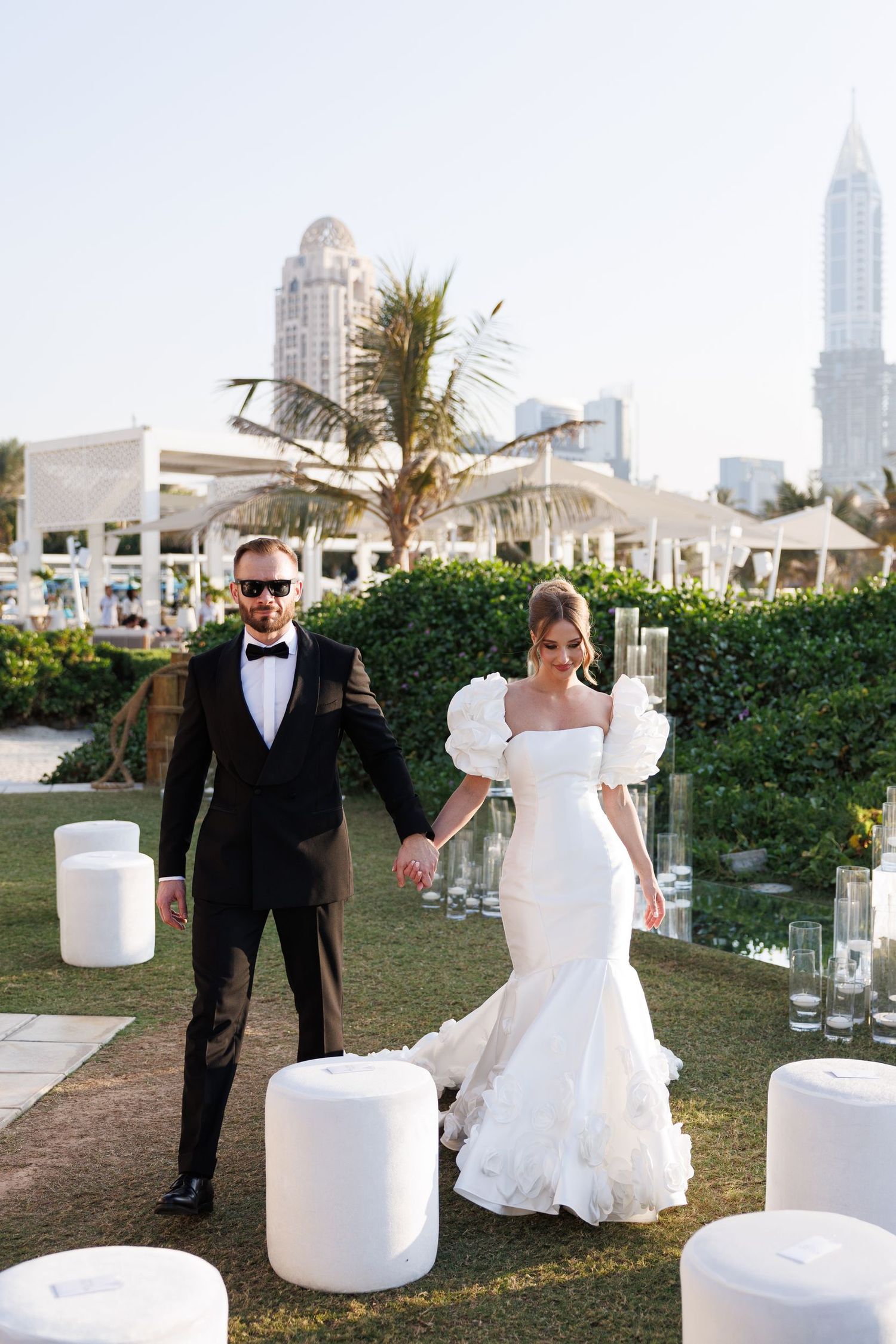 Wedding couple walking past elegant white tiered cakes displayed outdoors with city skyline in background.
