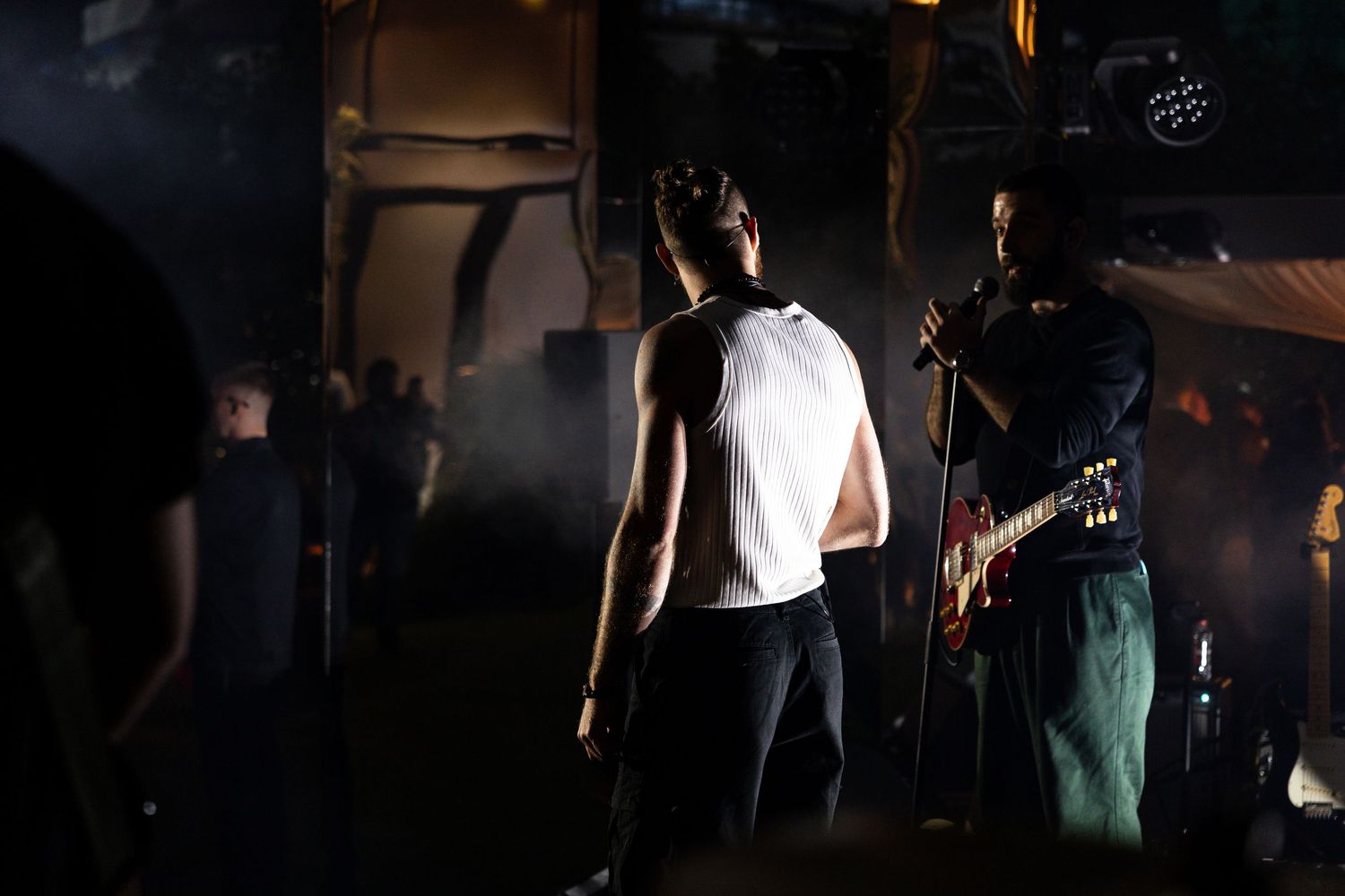 Musicians perform on stage at a dimly lit concert venue with guitars and microphones visible.