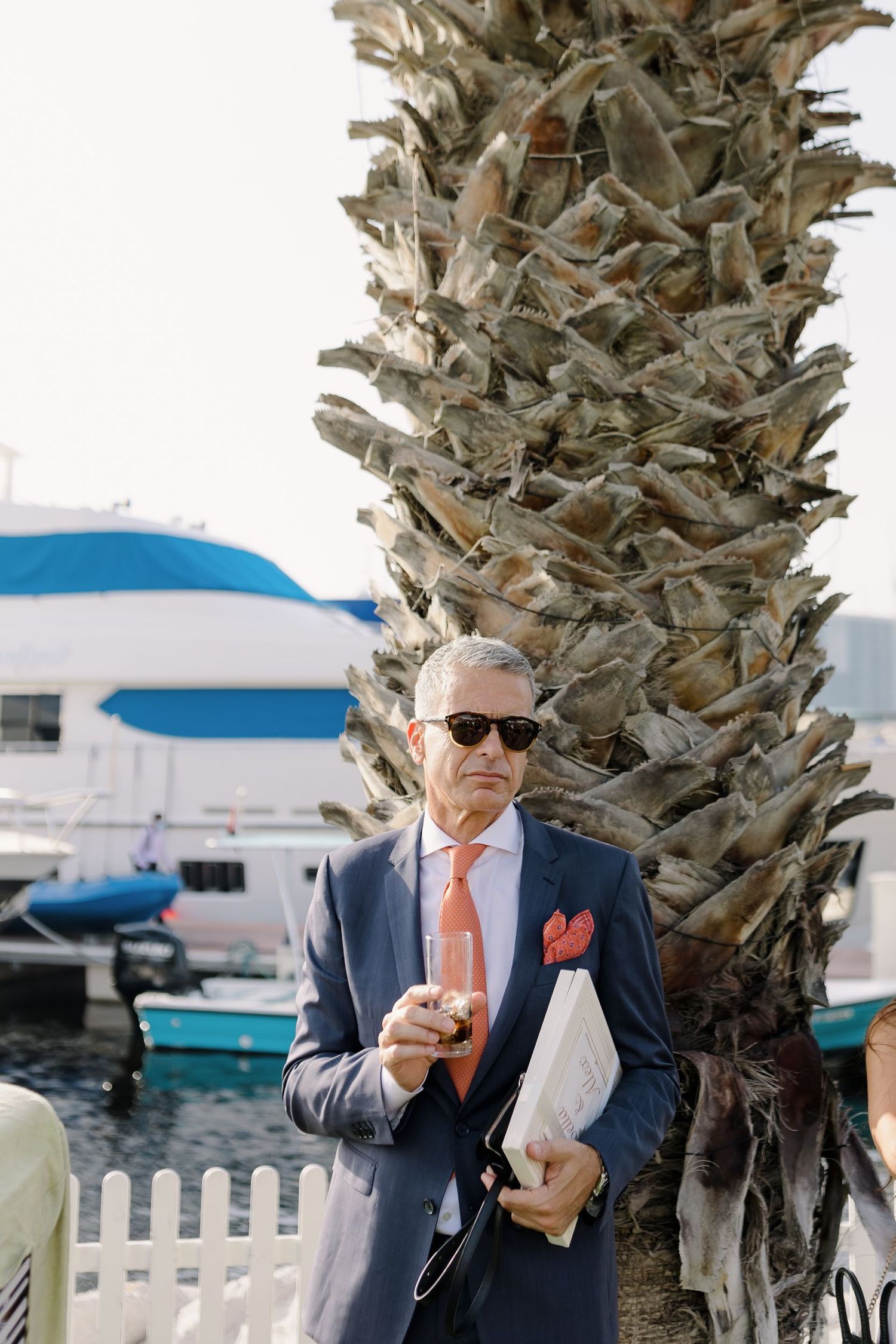 Person in a suit enjoying refreshments under palm trees at a waterfront venue.